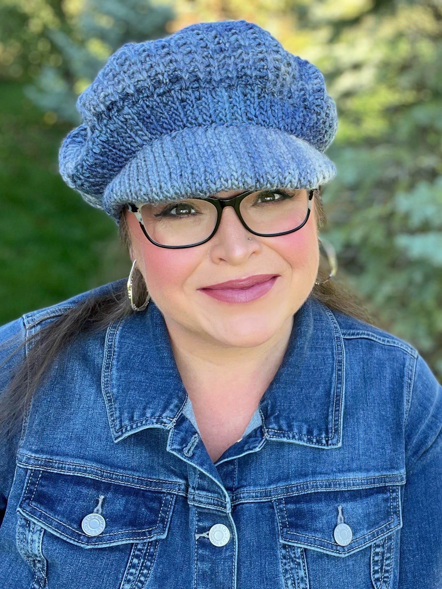 A woman smiles outdoors amid greenery, wearing the All About It Newsboy Crochet Hat (blue with a slip-stitch ribbed bill), glasses, hoop earrings, and a denim jacket.