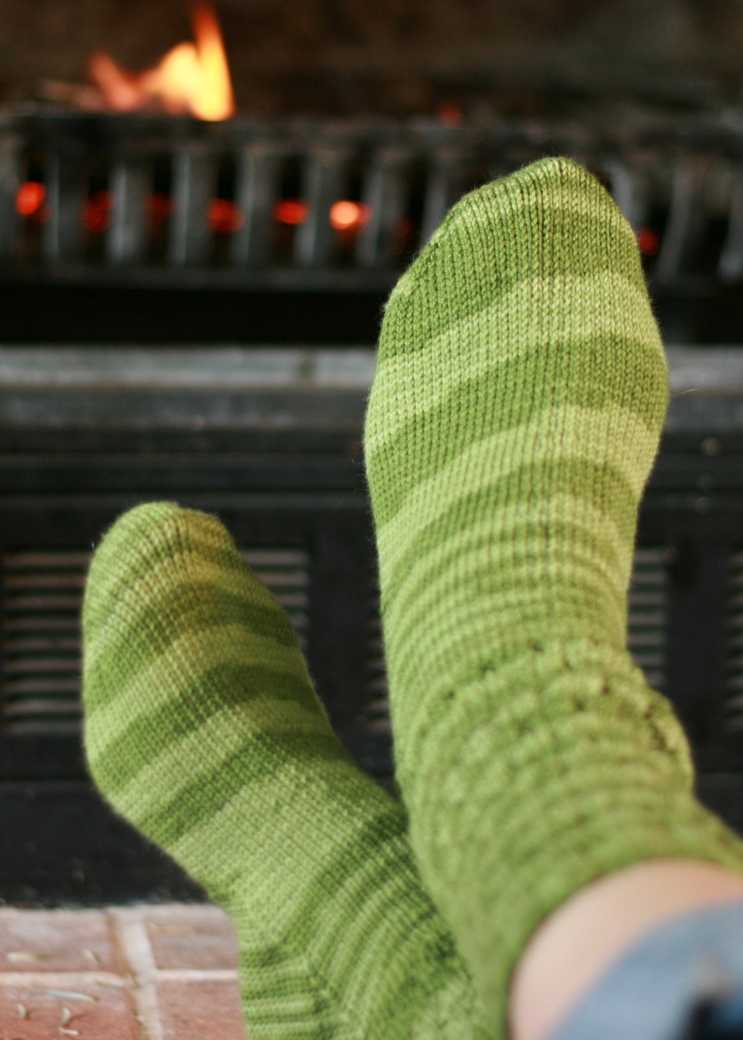 Someone relaxes with their feet up by a warm fireplace, wearing cozy socks they've made using the Marly Knit Socks Pattern.