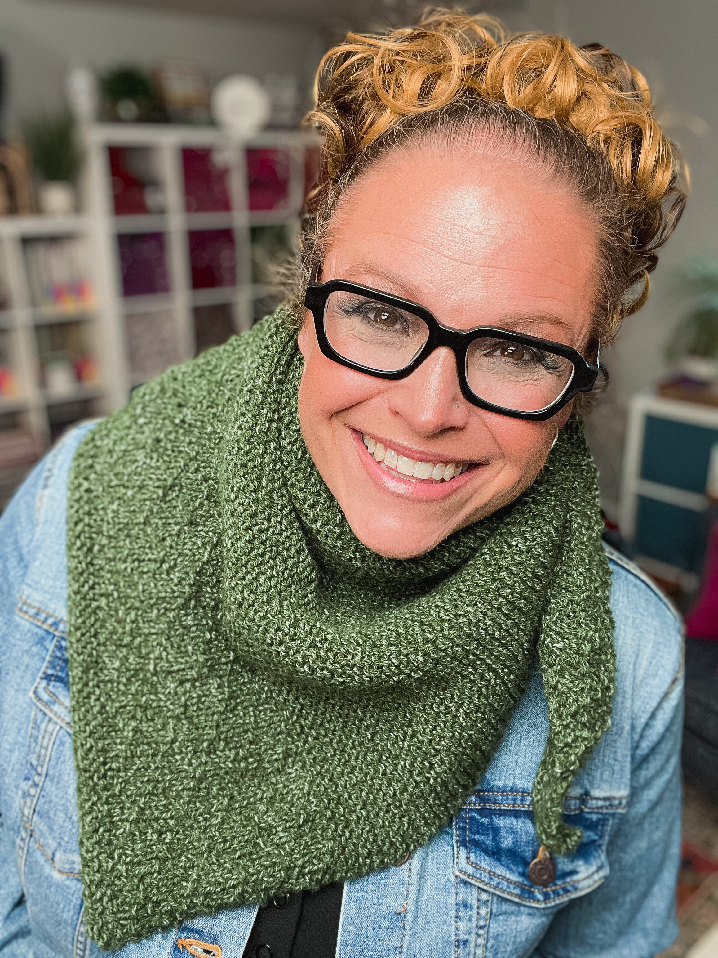 A smiling person with curly blond hair, black glasses, and a denim jacket models a green scarf made from the Peak Serenity Knit Shawl Pattern, standing indoors with bookshelves and decor in the background.