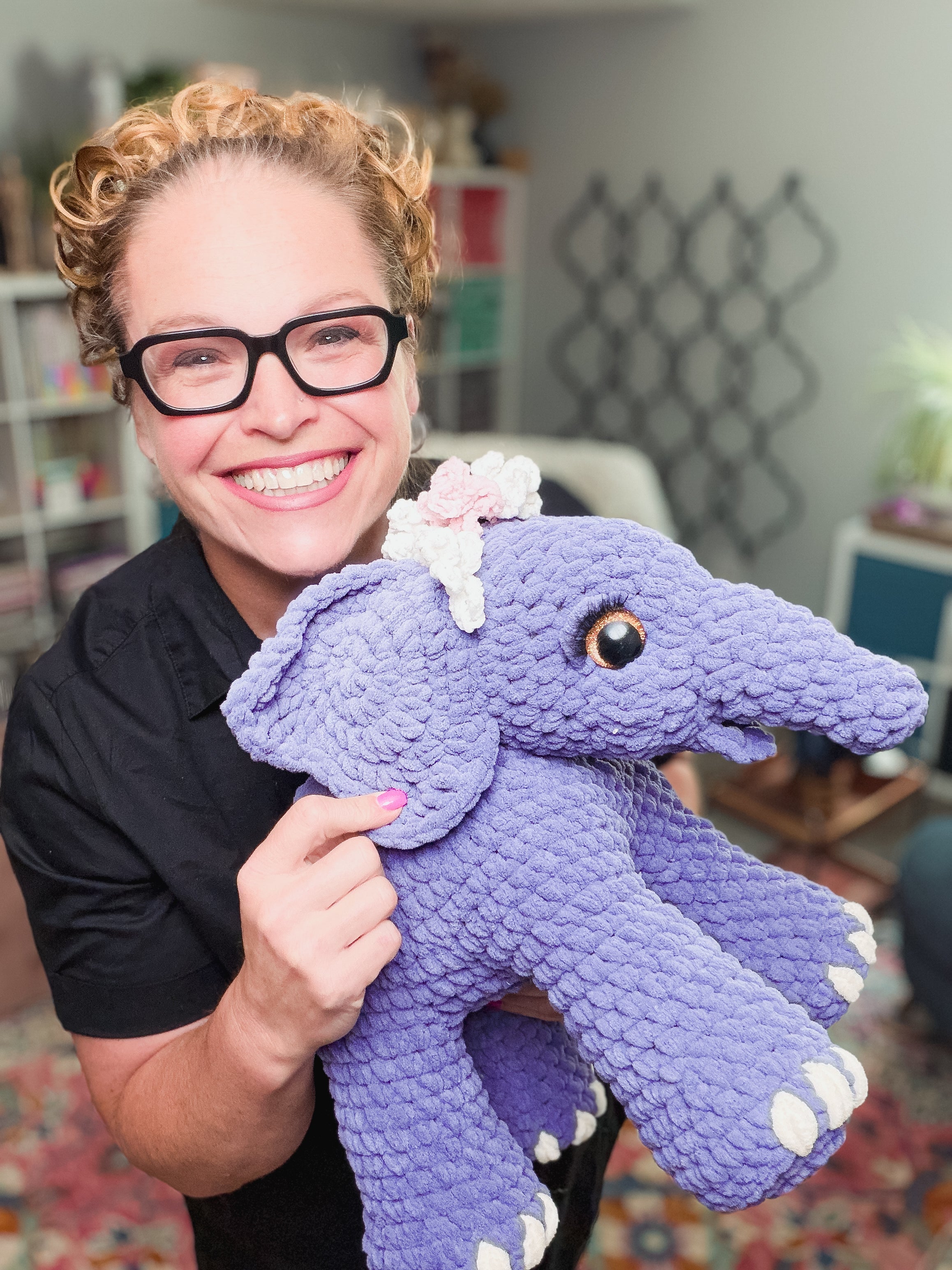 A smiling person with glasses holds Blossom the Crochet Amigurumi Elephant—a charming purple elephant with a flower crown—in a bright, cozy room.