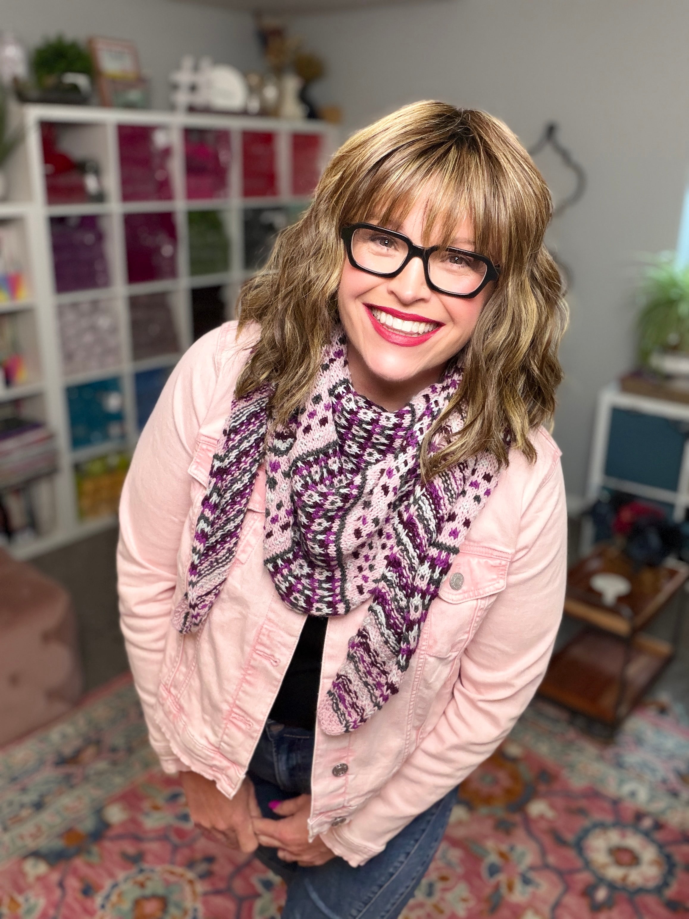 A smiling woman with wavy light brown hair, black glasses, a pink jacket, and a pink scarf stands in a cozy room displaying Anne Slip Stitch Mosaic Knit Shawl Pattern - Marly Bird.