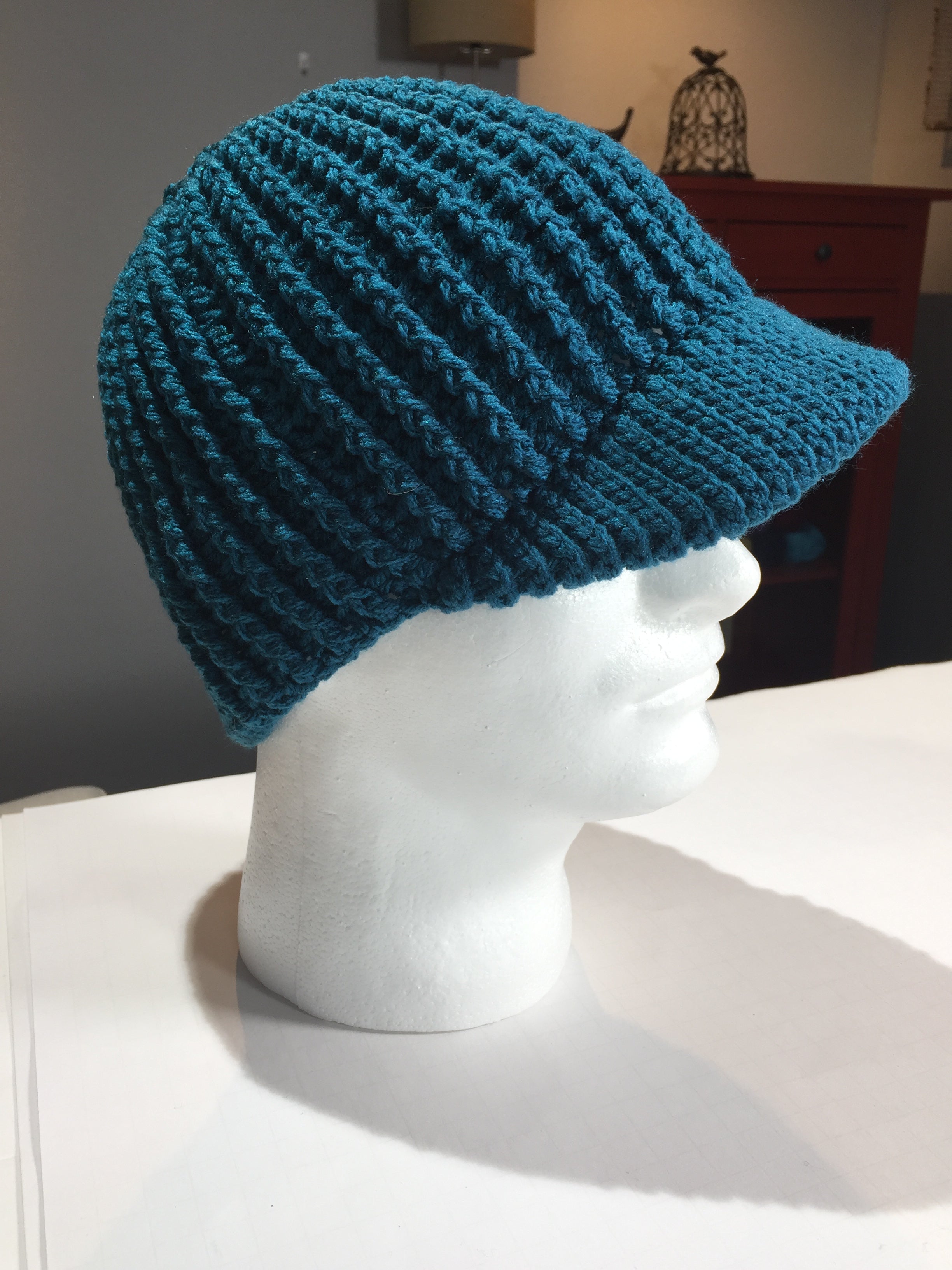 A white mannequin head showcases a teal hat made from the “Ribbed for Him & Her Crochet Hat Pattern PDF,” featuring ribbed crochet and a visor in a unisex beanie style, set on a white surface with softly blurred furniture and decor in the background.