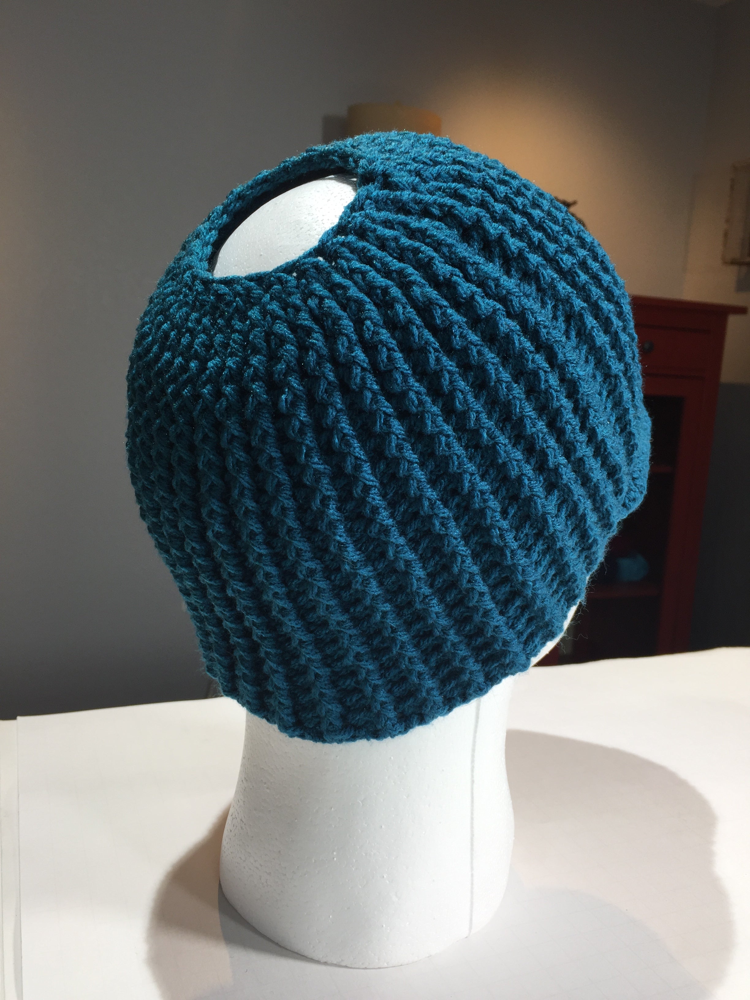 The white mannequin head displays a stylish unisex beanie made using the “Ribbed for Him & Her Crochet Hat Pattern PDF,” featuring a teal ribbed design with a large hole at the top, reminiscent of a classic ribbed crochet hat.