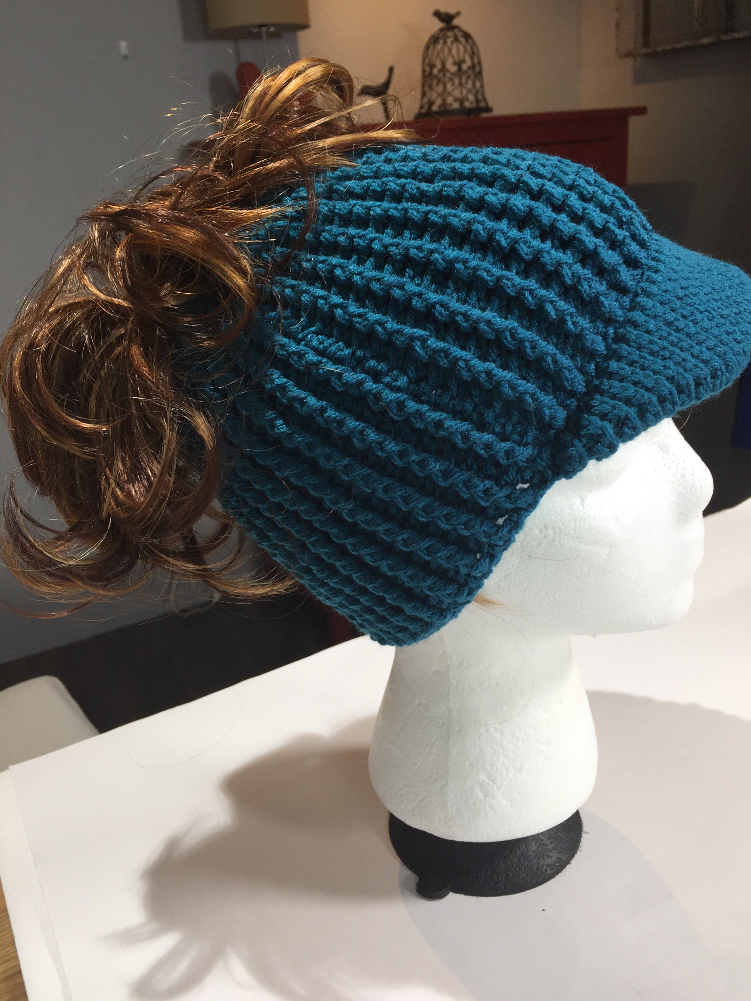 The Ribbed for Him & Her Crochet Hat Pattern PDF is shown in teal and features a visor, displayed on a white mannequin head. This unisex beanie includes an opening at the back, allowing a brown curly ponytail to stick out, combining practicality with style.