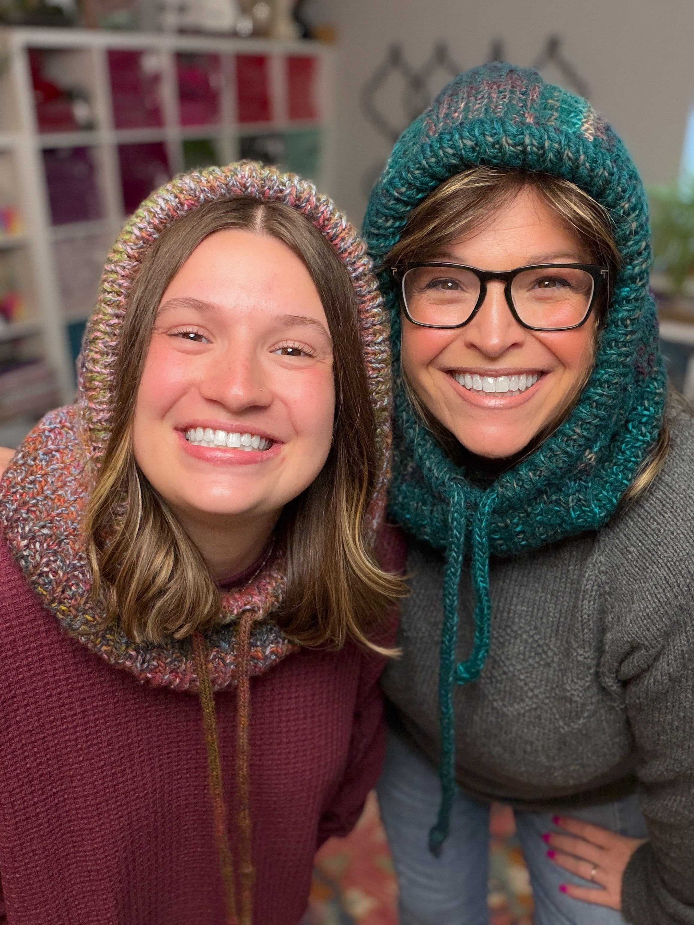 Two people smile indoors, each wearing a Cozy Sideline Knit Balaclava - PDF in multicolor and teal—ideal for showcasing your favorite knit pattern. Shelves of yarn fill the background as they pose in sweaters.