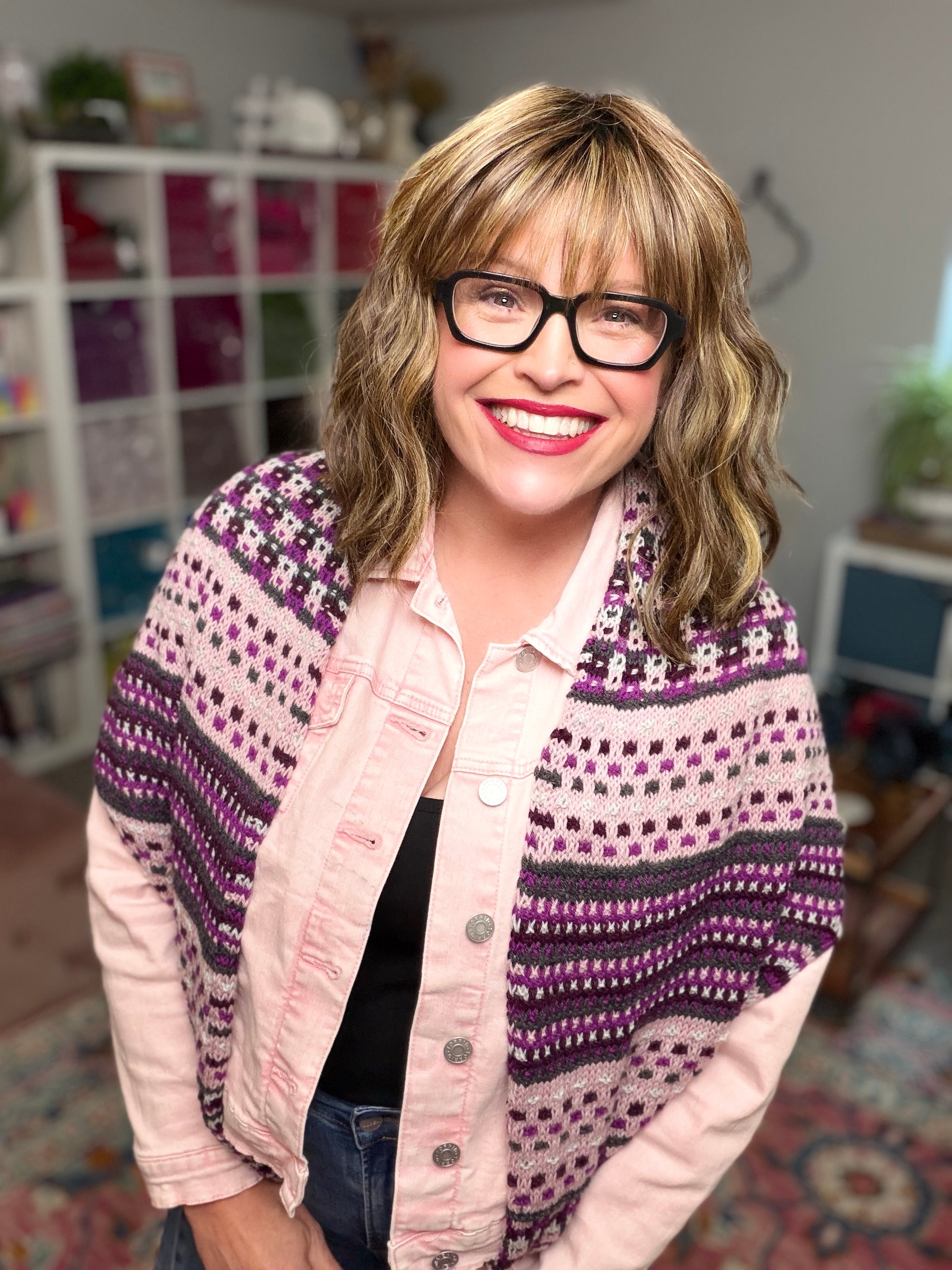 A smiling person with glasses and wavy hair wears a pink jacket and the Anne Slip Stitch Mosaic Knit Shawl Pattern - Marly Bird, standing in a cozy room with shelves and plants in the background.