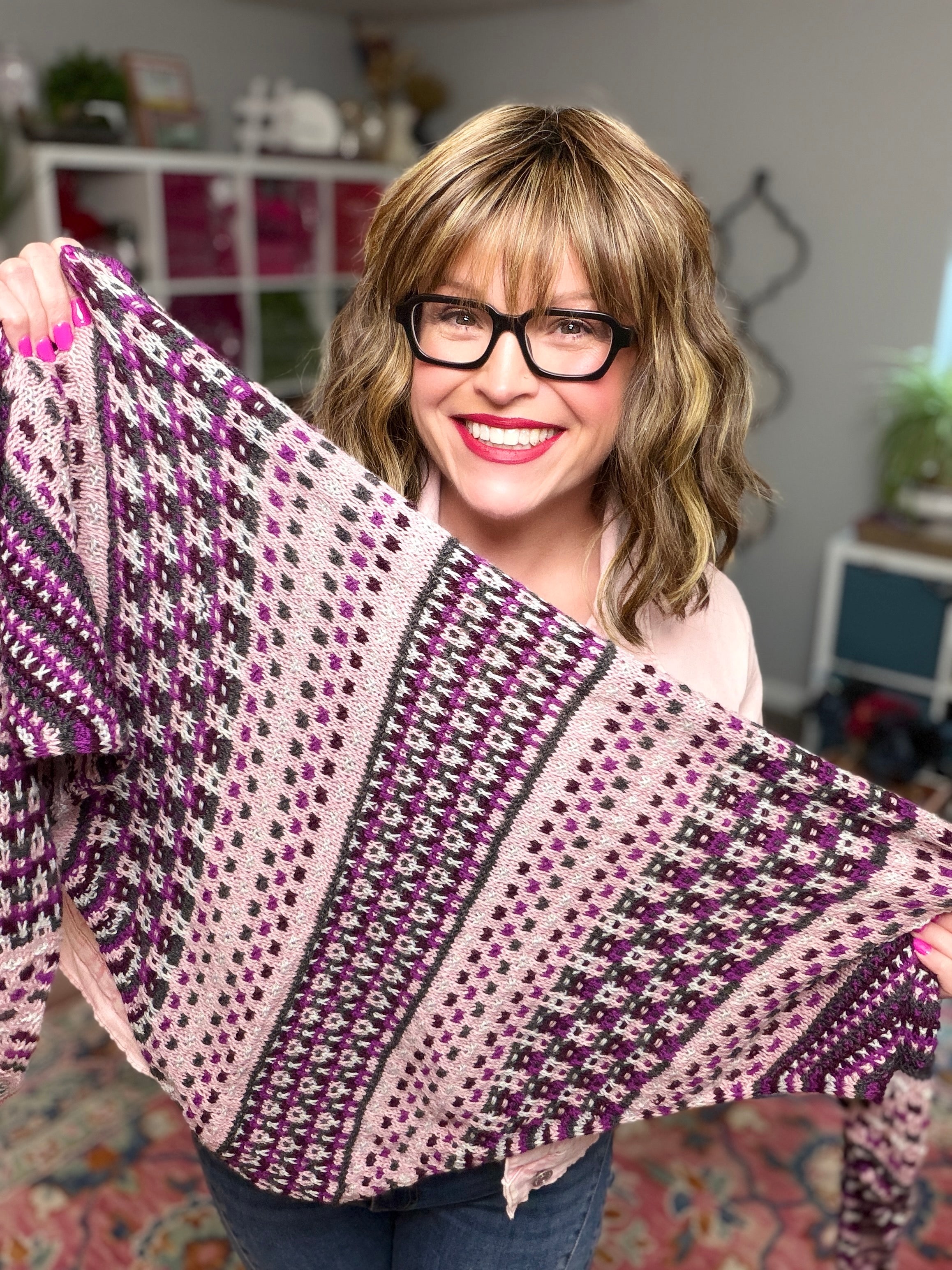 A smiling woman with glasses displays the Anne Slip Stitch Mosaic Knit Shawl Pattern - Marly Bird in pink and purple merino wool, standing in a cozy room with bookshelves and plants.