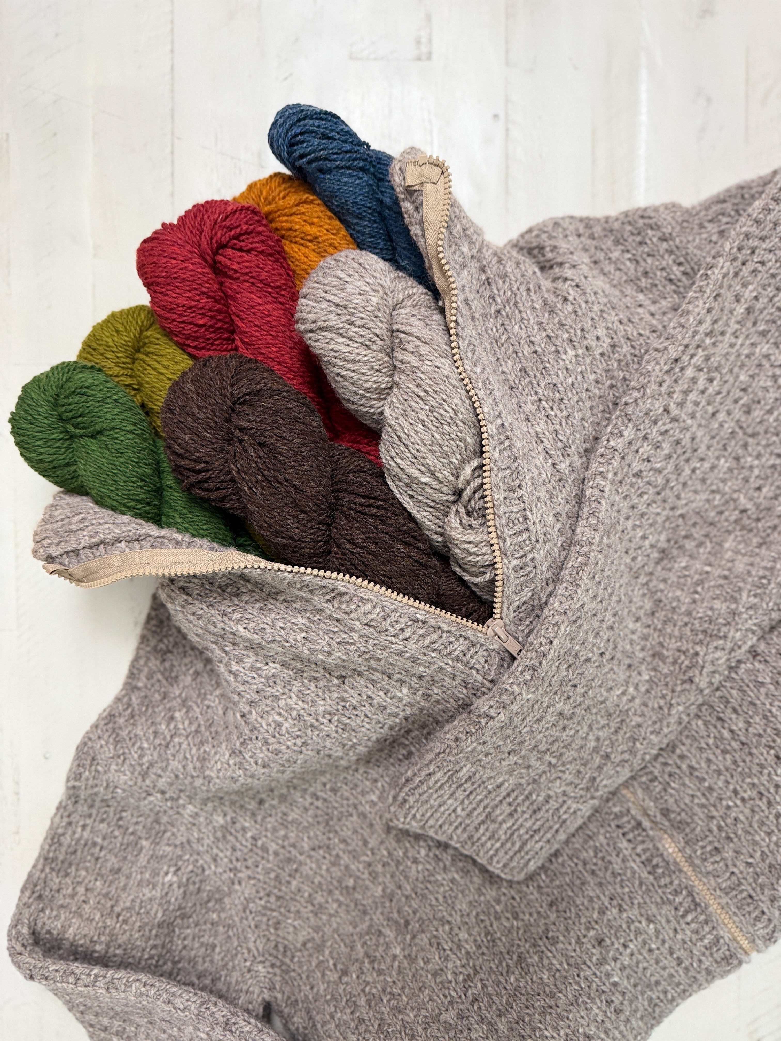 Adam’s Ribbed Zipper Knit Cardigan Pattern is displayed in beige, unzipped to reveal six skeins of yarn—red, green, yellow, blue, brown, and gray—arranged side by side on a light wooden surface. This unisex pattern is designed for inclusive sizing and effortless style.