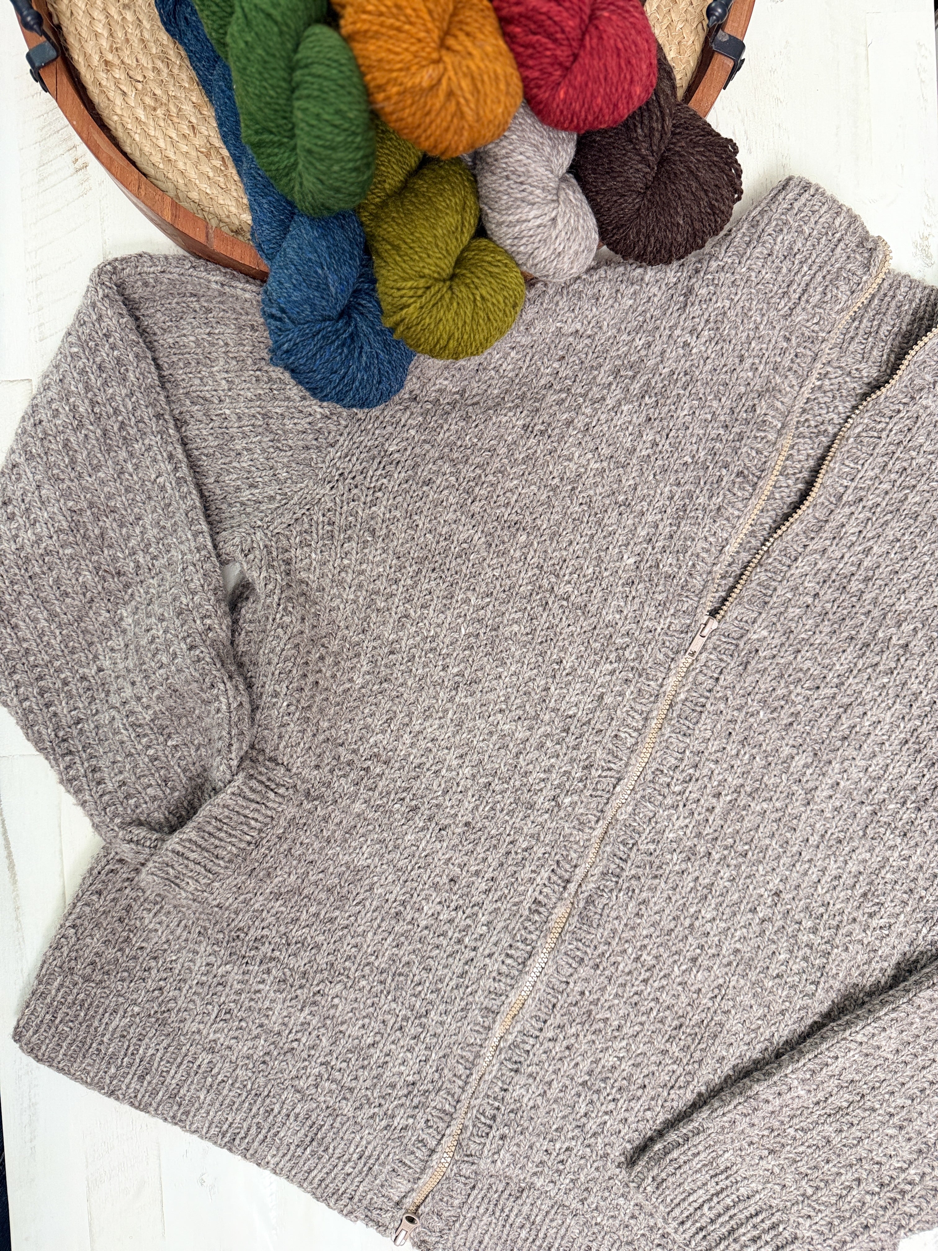 Adam’s Ribbed Zipper Knit Cardigan Pattern is shown laid flat in a textured, light gray design, next to a basket filled with skeins of yarn in blue, green, yellow, orange, gray, and brown on a white surface. This unisex pattern blends comfort and style for everyone.