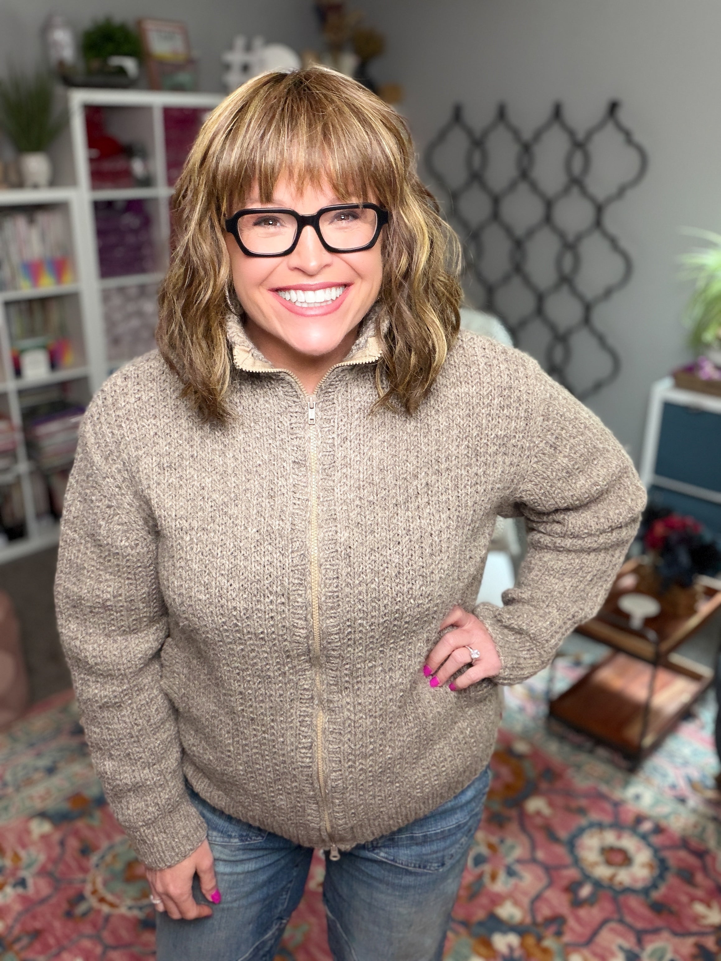 A smiling woman with glasses and shoulder-length light brown hair stands indoors, wearing a cardigan made using the Adam’s Ribbed Zipper Knit Cardigan Pattern and a pair of jeans. Shelves filled with books and decor can be seen in the background.