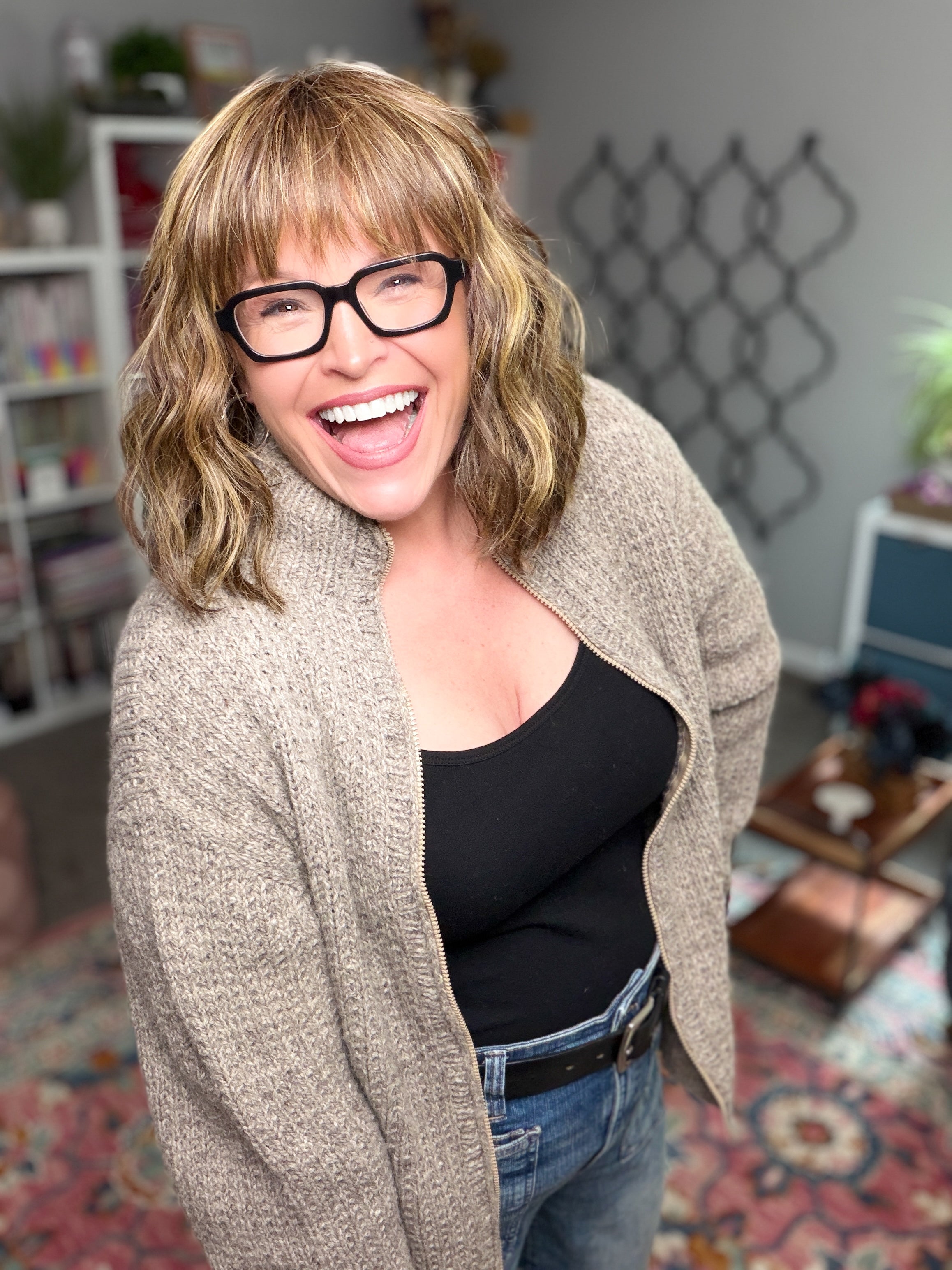 A woman with wavy blonde hair, glasses, and a big smile stands in a cozy, colorful room filled with shelves, plants, and a patterned rug. She wears a black top layered with the Adam’s Ribbed Zipper Knit Cardigan—crafted from an inclusive pattern designed to ensure comfort and style for everyone.