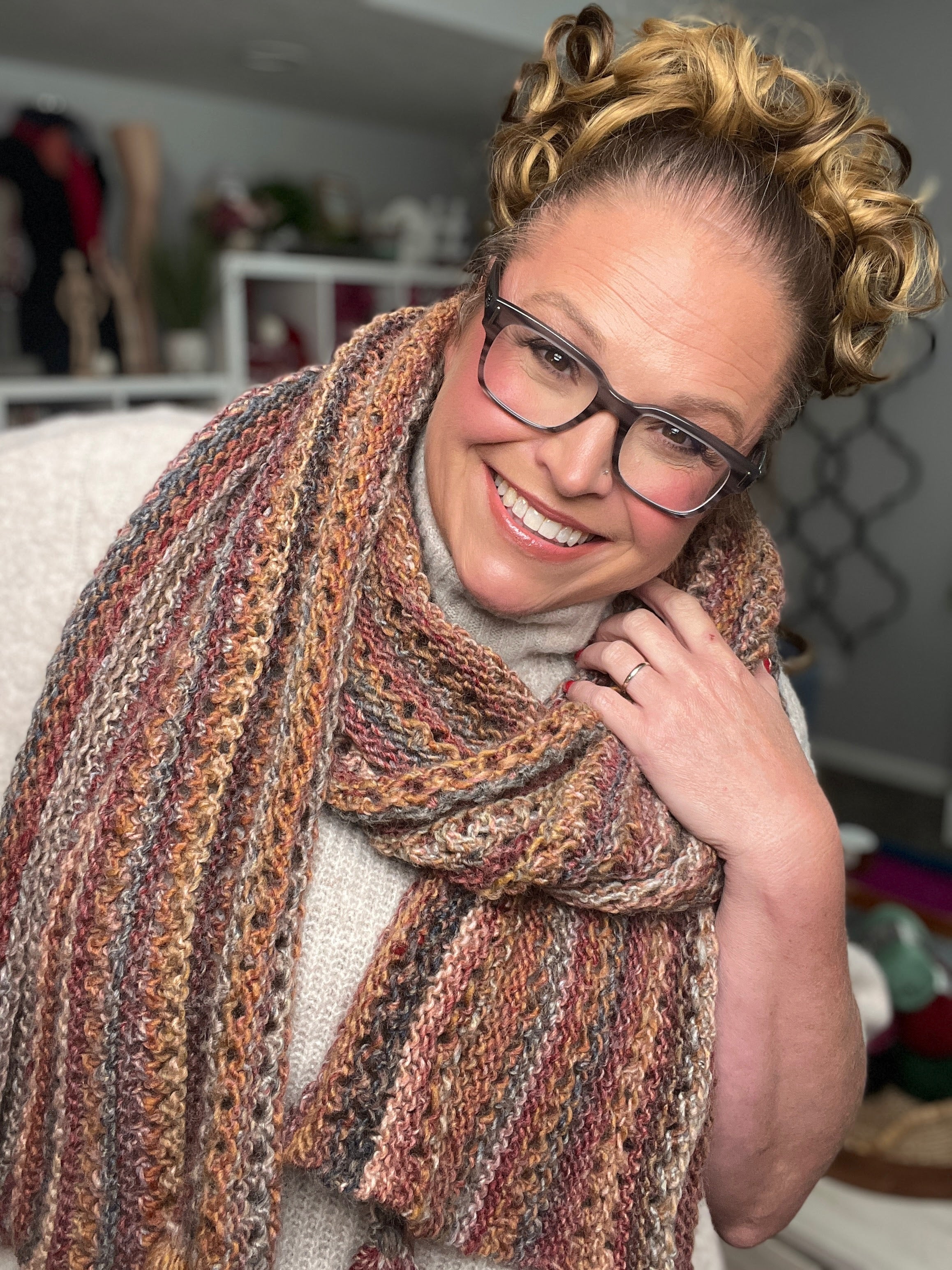 A smiling woman with curly blonde hair, glasses, and rosy cheeks models the Almond Brittle Knit Wrap Pattern—a chunky, self-striping bulky lace scarf wrap with tassels—indoors, wearing a beige top. Shelves and home decor appear in the background.