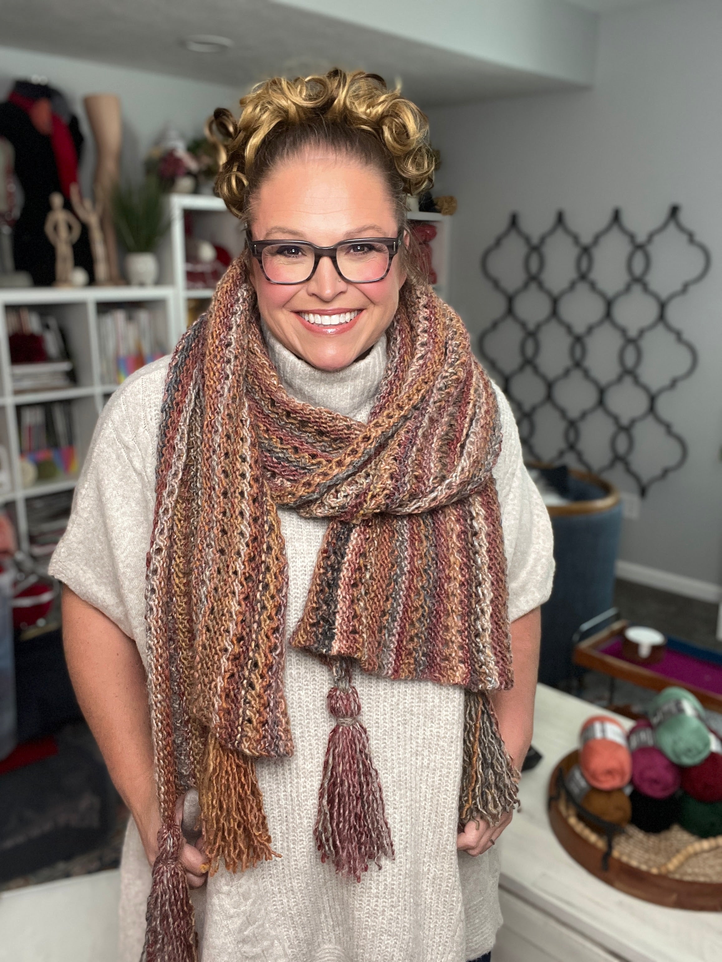 A smiling person with glasses and curly hair models the Almond Brittle Knit Wrap Pattern— a bulky lace knit scarf wrap with tassels, worn over a light sweater, with shelves of yarn and a decorative lattice on a gray wall behind.
