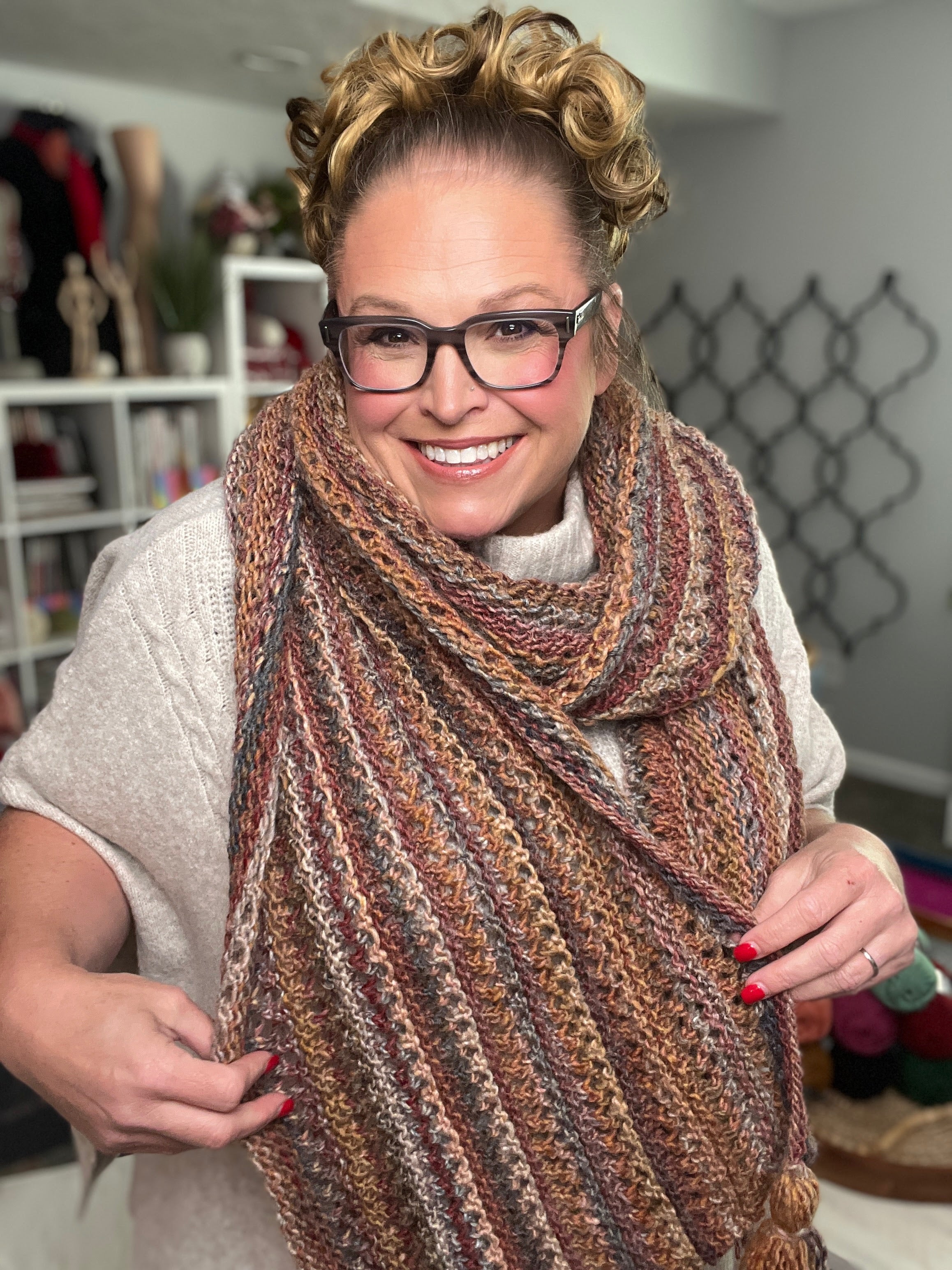A smiling woman with glasses and curly hair wears the Almond Brittle Knit Wrap Pattern— a bulky lace knit scarf wrap with tassels—over a beige sweater, standing in a cozy room with yarn and shelves in the background.