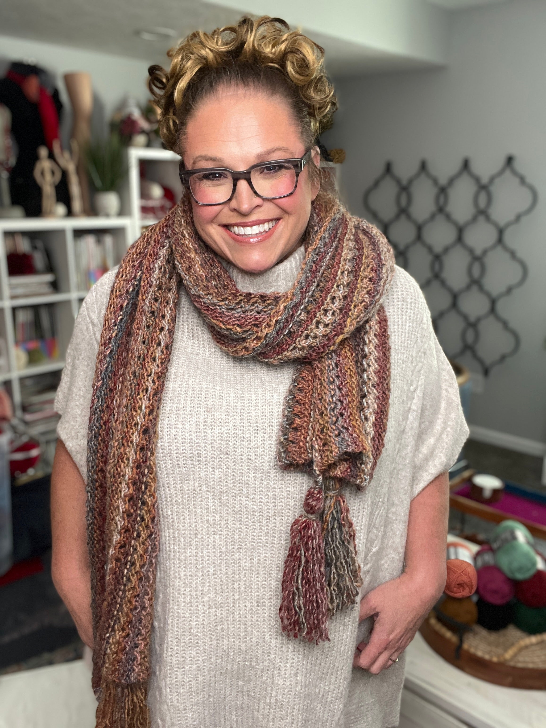A smiling woman with curly hair and glasses wears a beige sweater and the Almond Brittle Knit Wrap, a bulky lace knit scarf with tassels. She stands in a cozy room filled with bookshelves, yarn, and craft supplies.