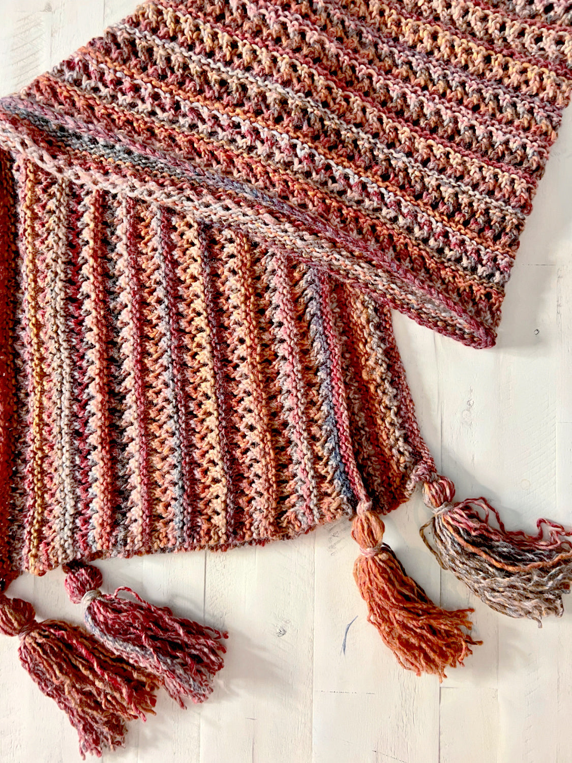 The Almond Brittle Knit Wrap Pattern is a bulky lace knit scarf PDF with tassels, shown in variegated pink, red, and beige tones and displayed on a light wooden surface.