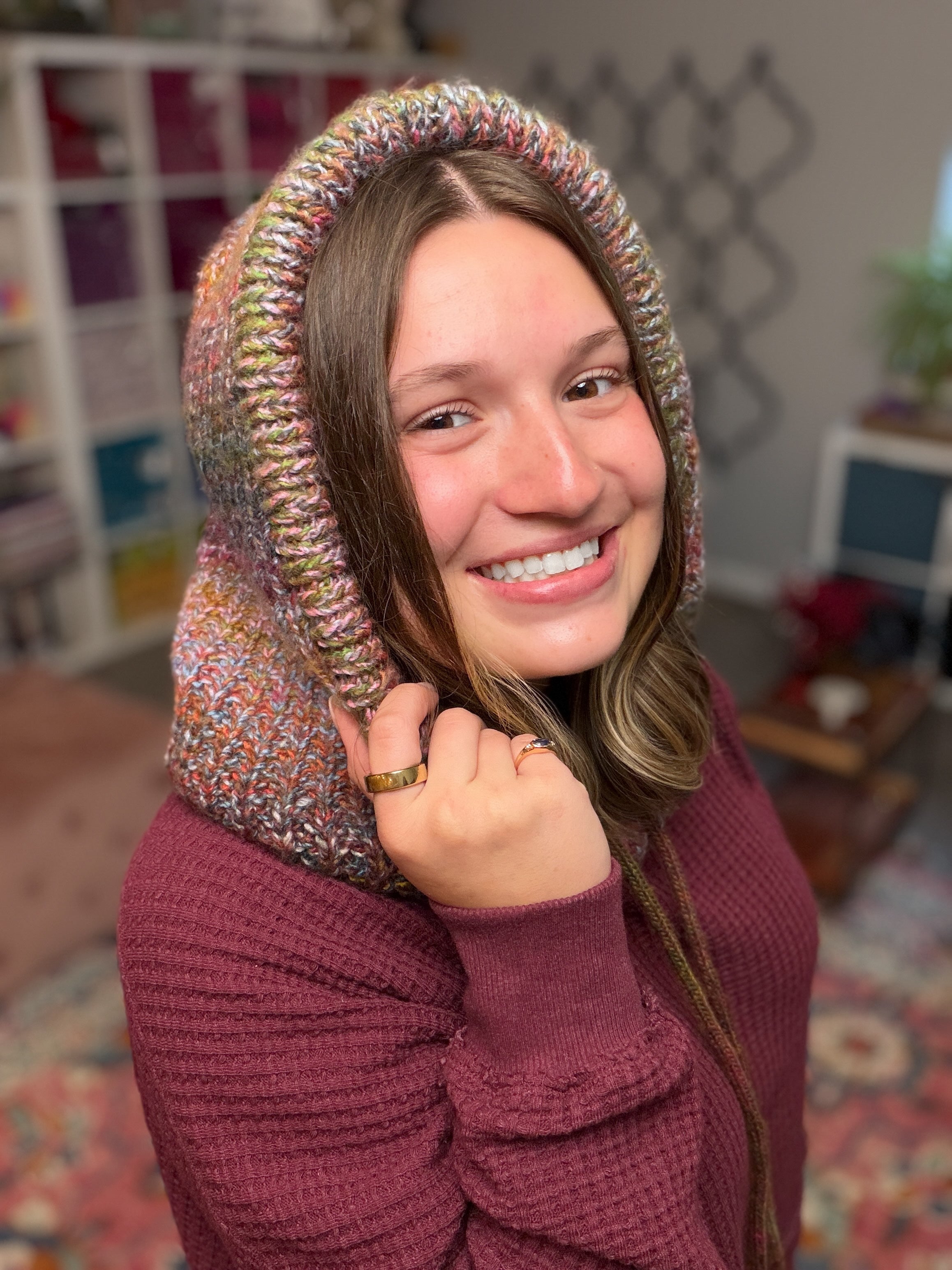 A smiling woman in a maroon sweater holds up the Cozy Sideline Knit Balaclava - PDF in a cozy, well-lit room with shelves and a patterned rug in the background.