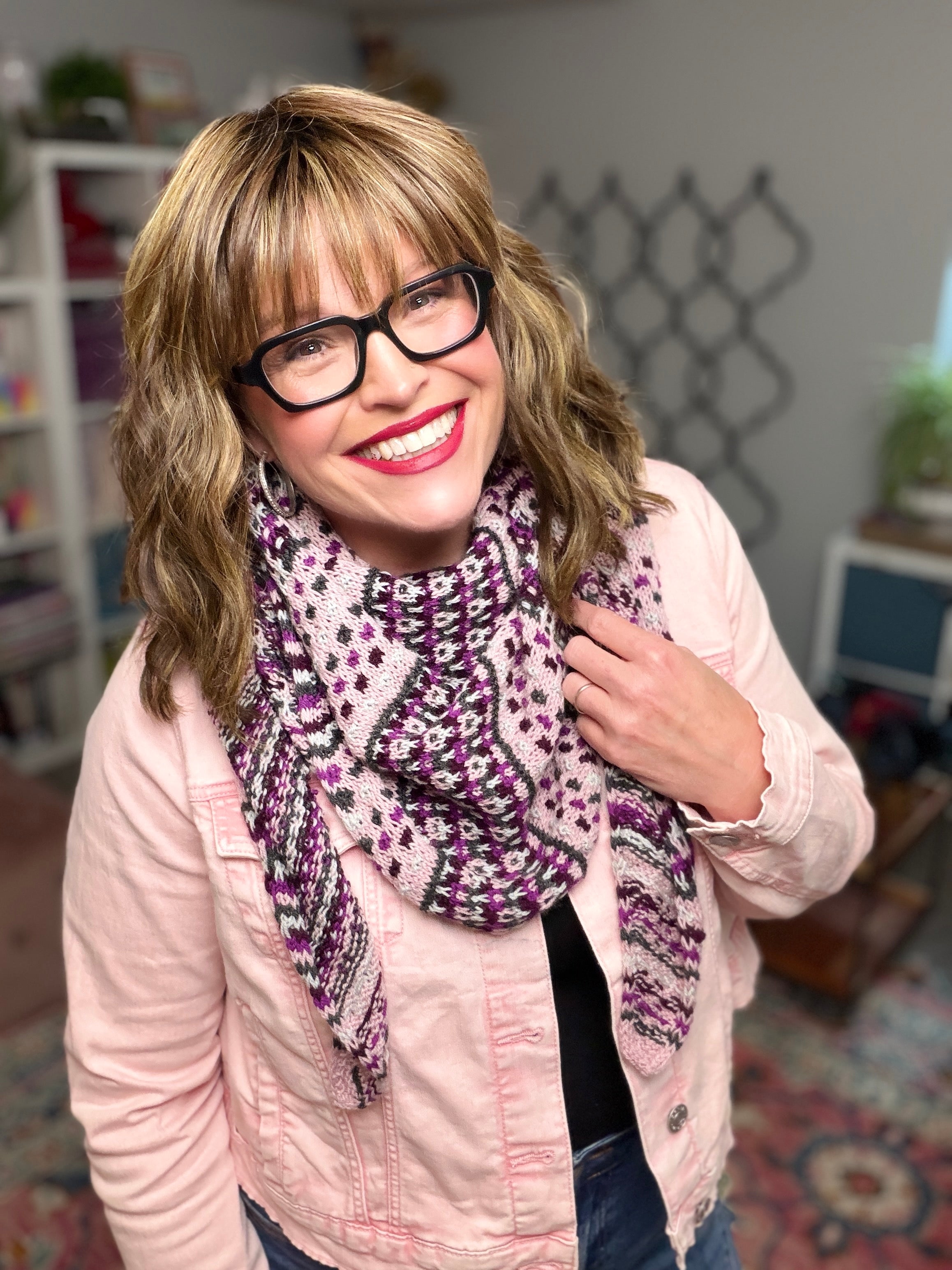 A smiling woman with wavy blonde hair wears black glasses, a pink jacket, and the Anne Slip Stitch Mosaic Knit Shawl Pattern - Marly Bird, standing indoors with shelves and plants behind her.