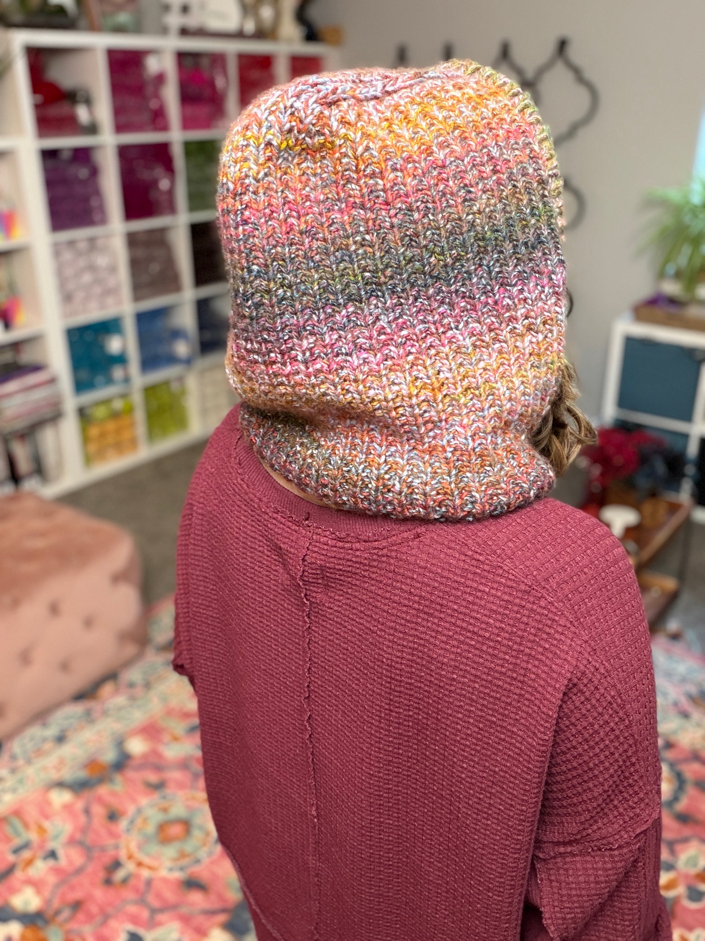 A person wearing the Cozy Sideline Knit Balaclava - PDF and a maroon sweater stands in a cozy room with cubed shelves of worsted weight yarn, a pink ottoman, and a patterned rug.