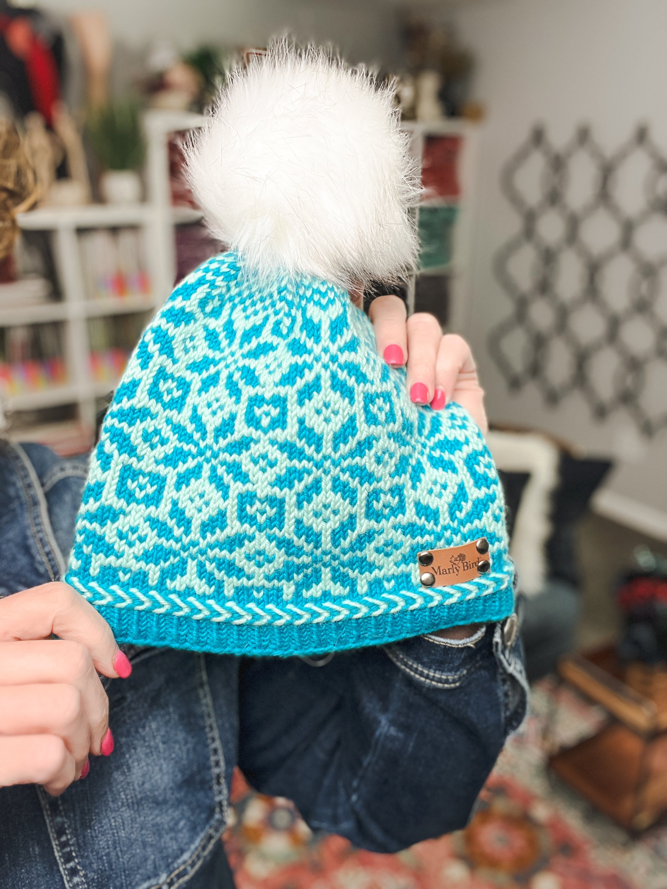 A person with pink nails holds the Chill Out Winter Fair Isle Hat—a blue and white knit winter hat with a white fluffy pom-pom on top, featuring an eye-catching colorwork pattern and a brown leather patch on the brim.