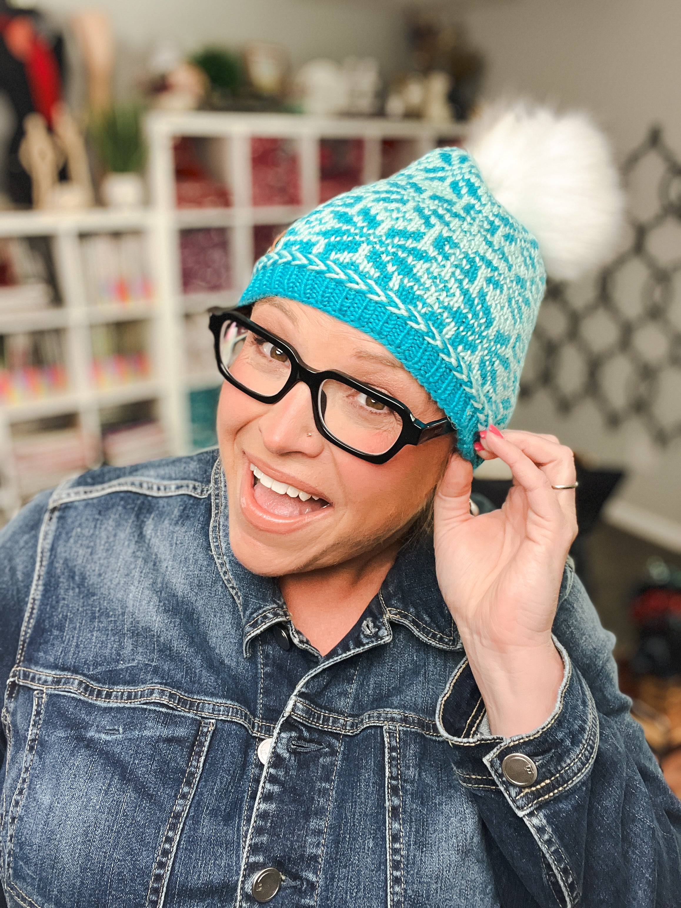 A smiling person wearing black glasses and a denim jacket poses indoors with shelves and yarn in the background, showcasing the Chill Out Winter Fair Isle Hat - PDF, a blue patterned knit hat with a white pom-pom.