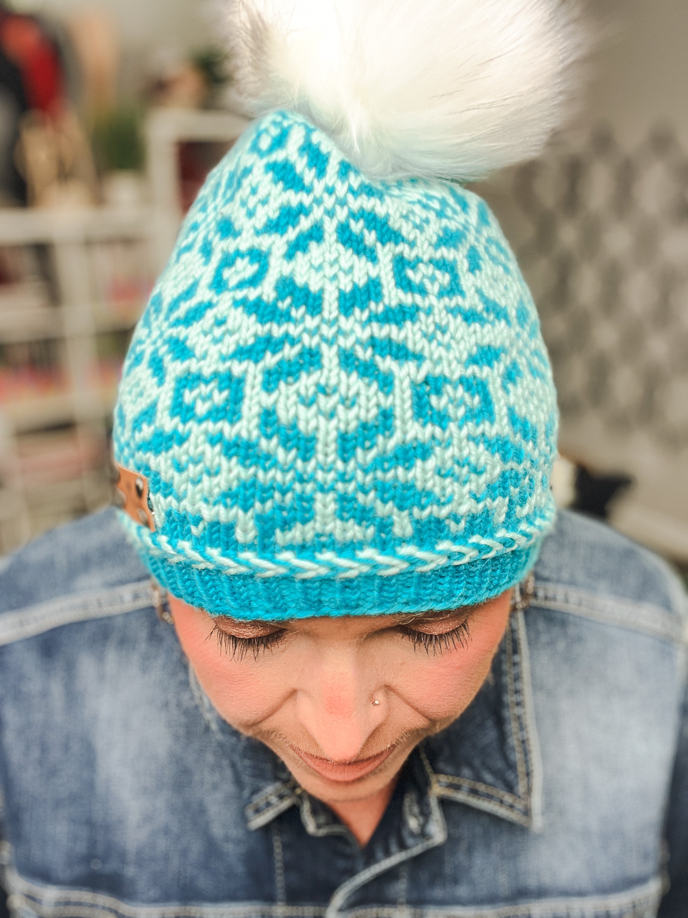 A person wearing the Chill Out Winter Fair Isle Hat, a blue patterned knit hat with a white pom-pom, looks down. They have a nose piercing and are dressed in a denim jacket, with the background blurred.