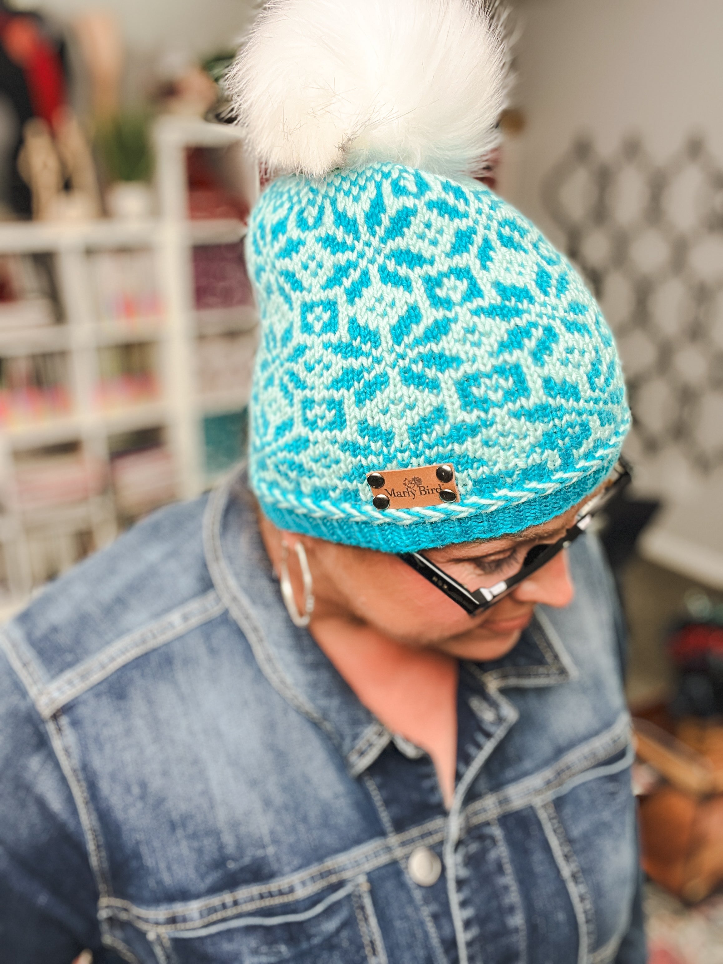 A person wearing glasses and a denim jacket looks downward indoors, modeling the Chill Out Winter Fair Isle Hat – PDF: a blue knit hat with a white pom-pom and intricate colorwork pattern, while shelves and decor are blurred in the background.