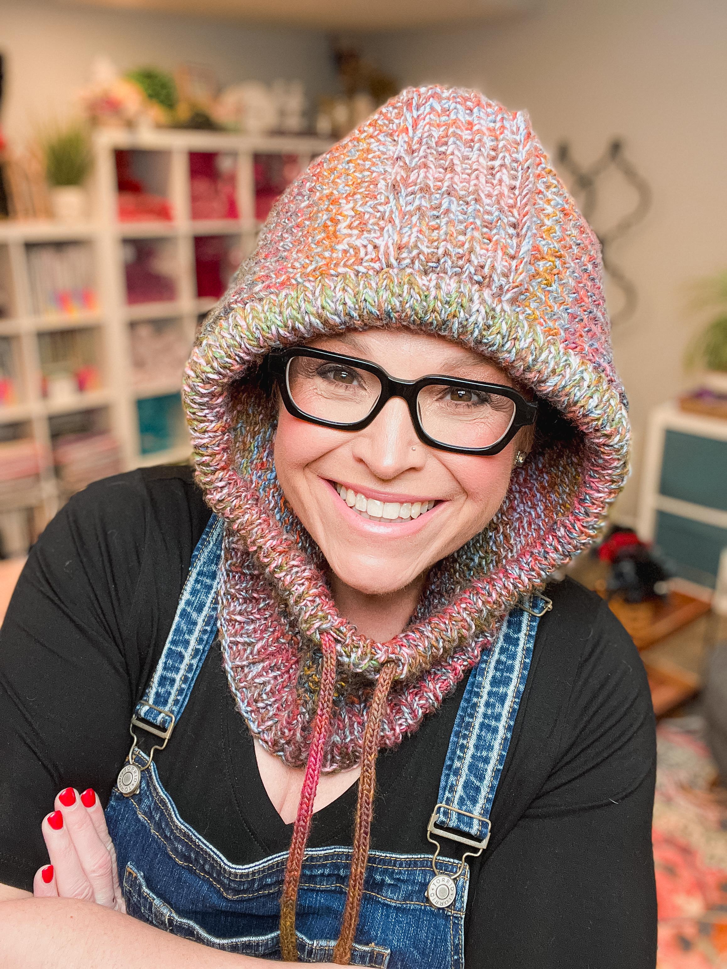 A smiling woman wearing a chunky hand-knit multicolor hooded cowl hat with black glasses, a black shirt, and blue denim overalls, set against a cozy, softly blurred craft room background.