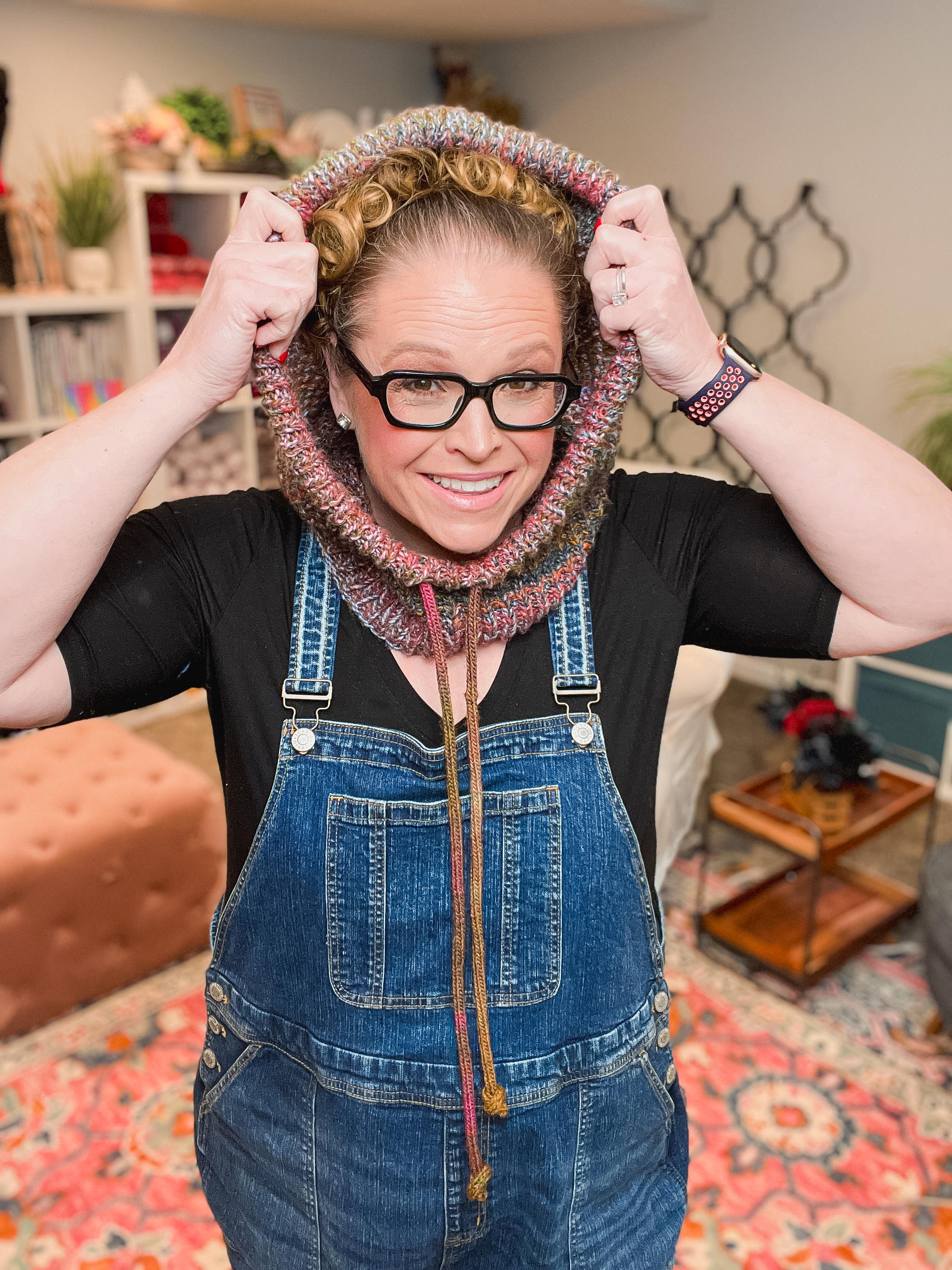 A woman wearing a chunky knit cowl neck warmer in a multicolor pattern, styled with a black t-shirt and blue denim overalls, holding the top of the cowl with both hands in a cozy indoor setting.