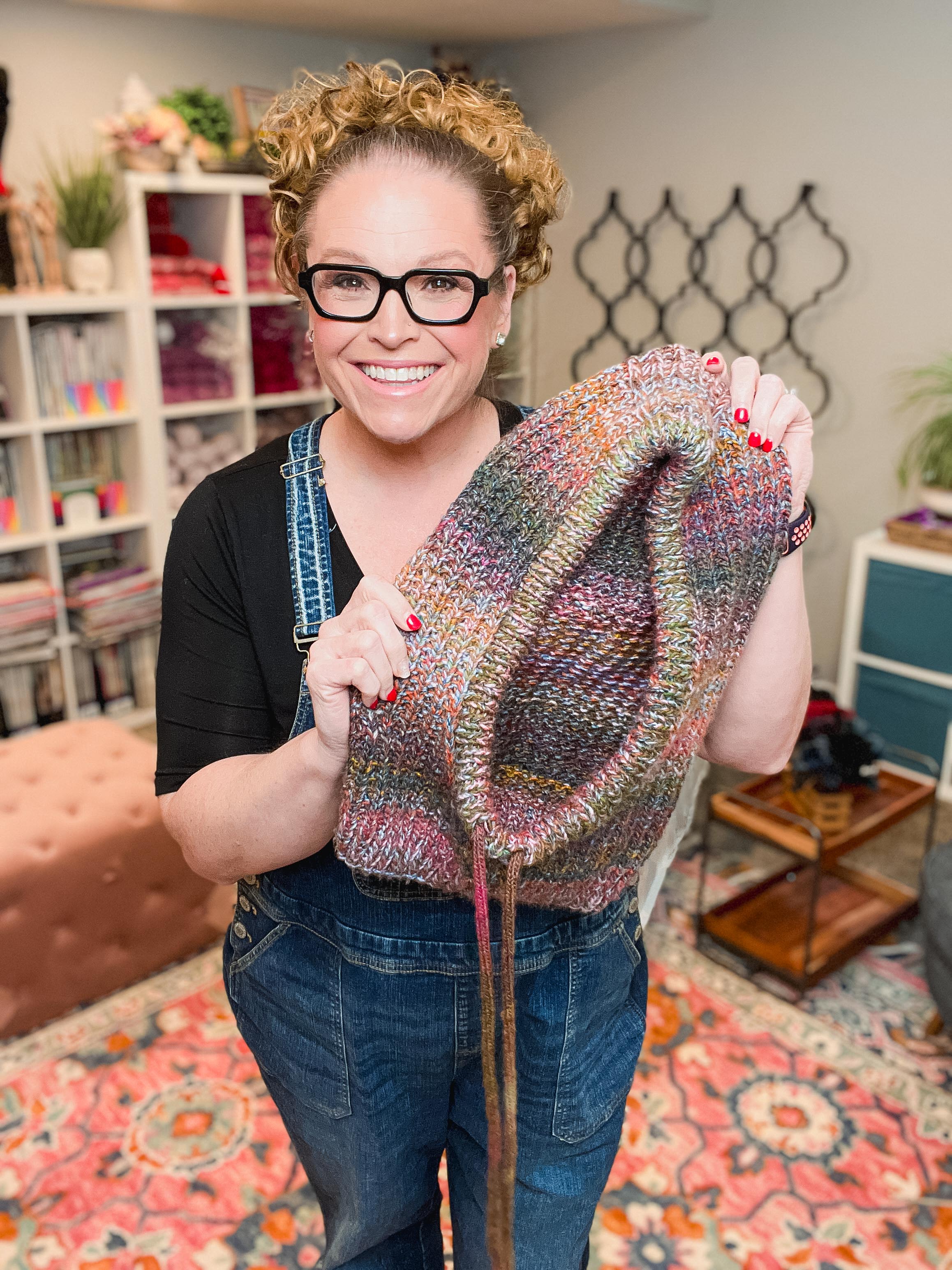 A smiling woman with curly blonde hair and black glasses holds up a large, hand-knit bag made from a variegated yarn blend of pink, blue, green, and orange. She is wearing blue denim overalls in a cozy, colorful craft room setting.