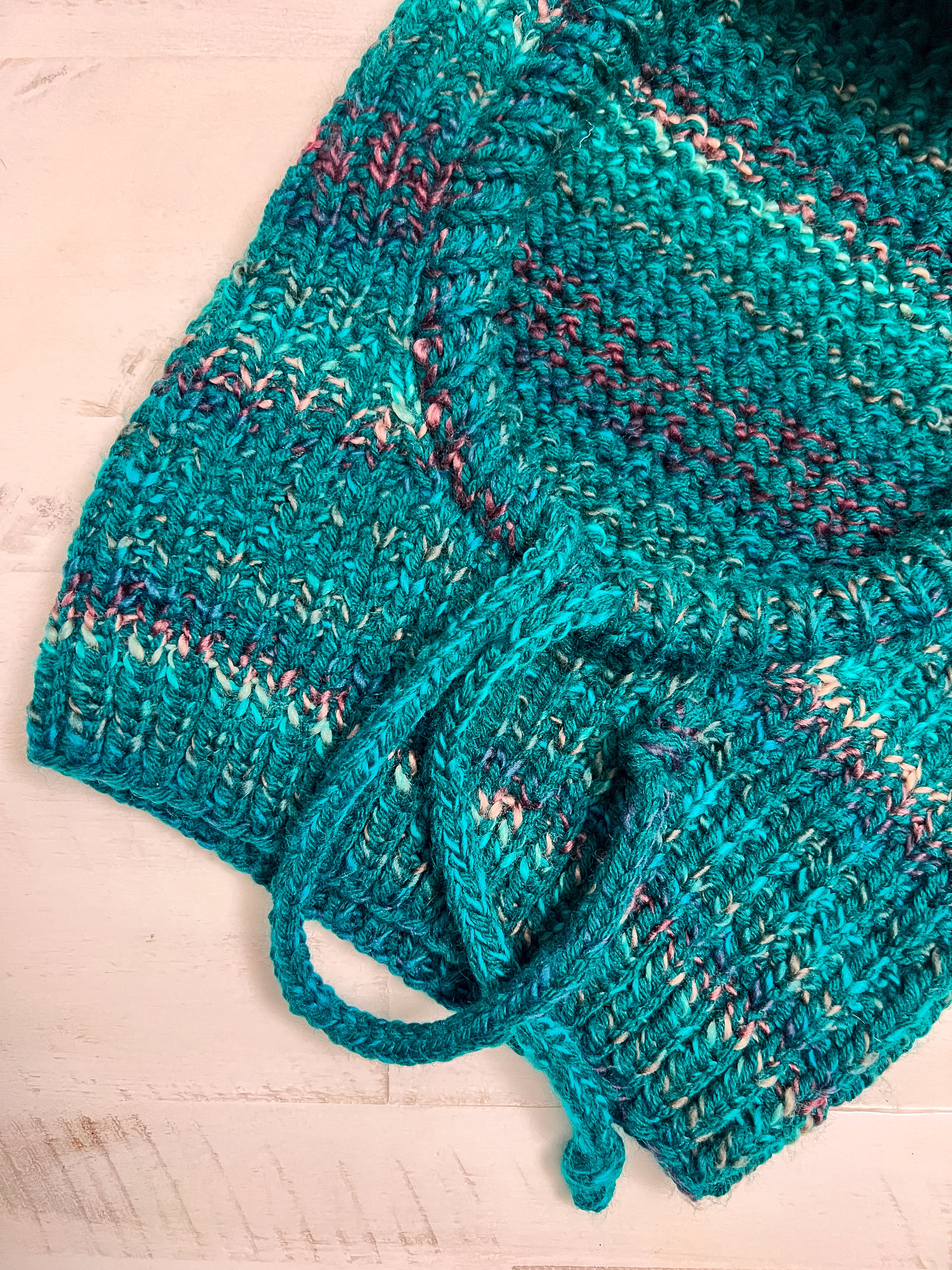 A close-up of the Cozy Sideline Knit Balaclava, crafted from turquoise worsted weight yarn with pink and white specks, showcases its textured tweed-like pattern and drawstring detail, displayed on a light wooden surface.
