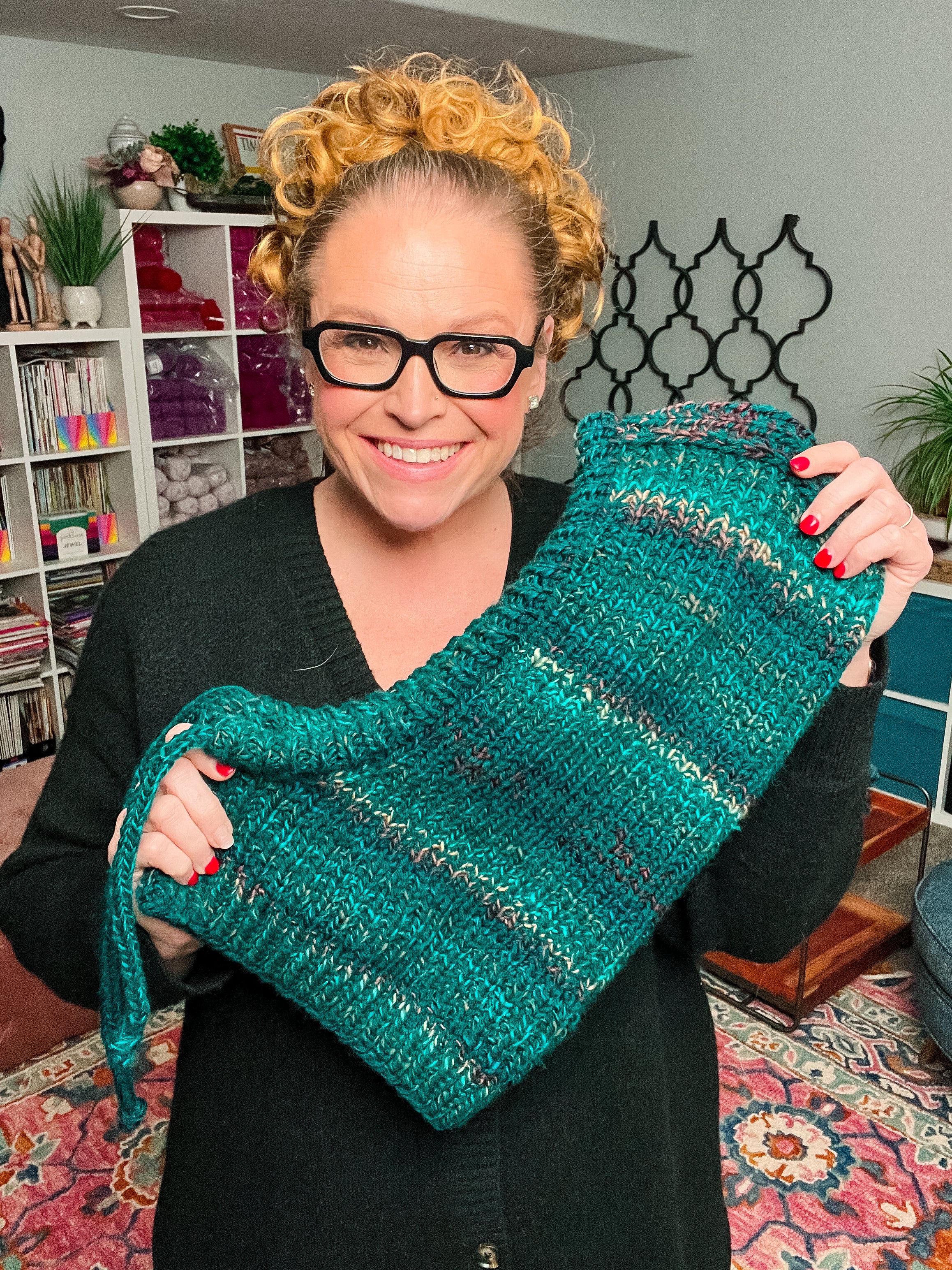 A smiling woman with curly hair and glasses holds up a thick, teal-blue Cozy Sideline Knit Balaclava—made from tweed-like fabric—in a cozy, colorful room filled with bookshelves and plants.