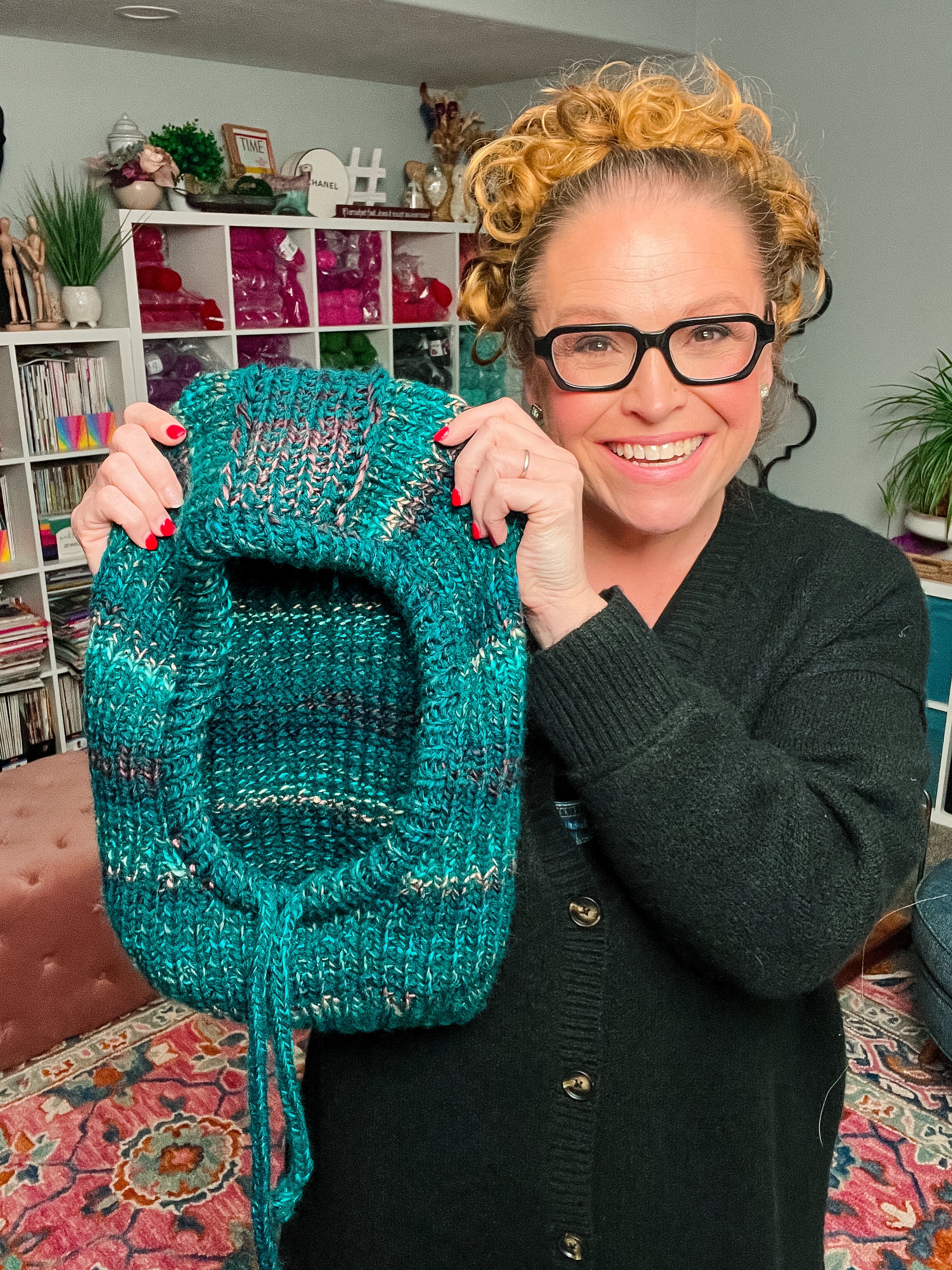 A smiling woman with glasses holds up the “Cozy Sideline Knit Balaclava”—a green hooded cowl crafted from worsted weight yarn—in a cozy, colorful room filled with yarn, books, and plants.