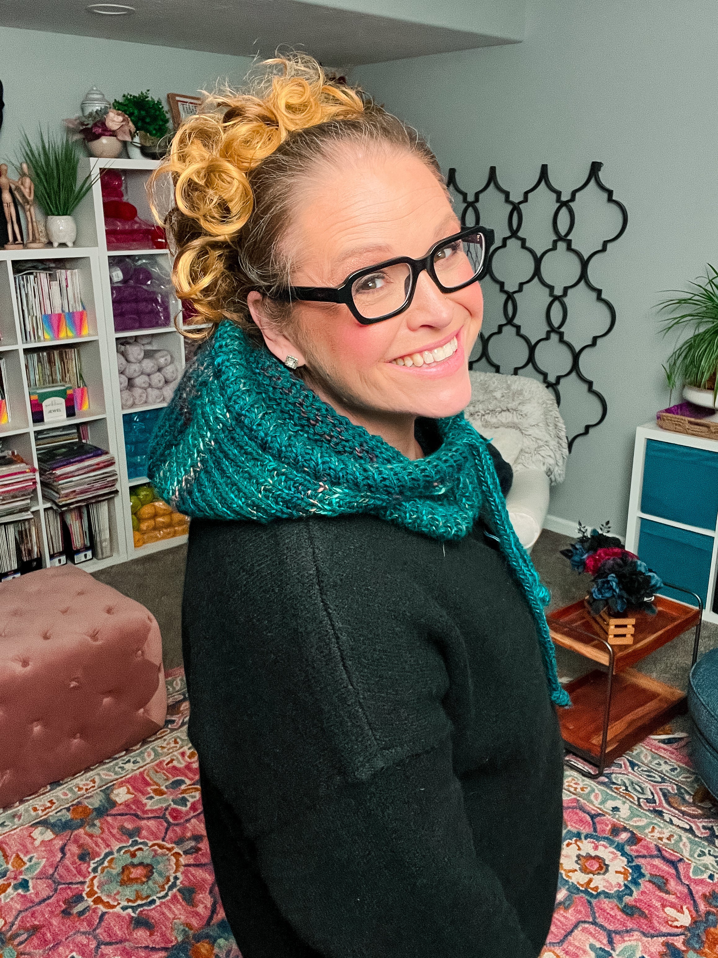 A smiling woman with glasses and curly hair wears a teal knit scarf in a cozy, colorful room filled with bookshelves, yarn, plants, and a patterned rug—an inviting space perfect for making your own Cozy Sideline Knit Balaclava - PDF, a customizable pattern designed for soft worsted weight yarn and the perfect fit.