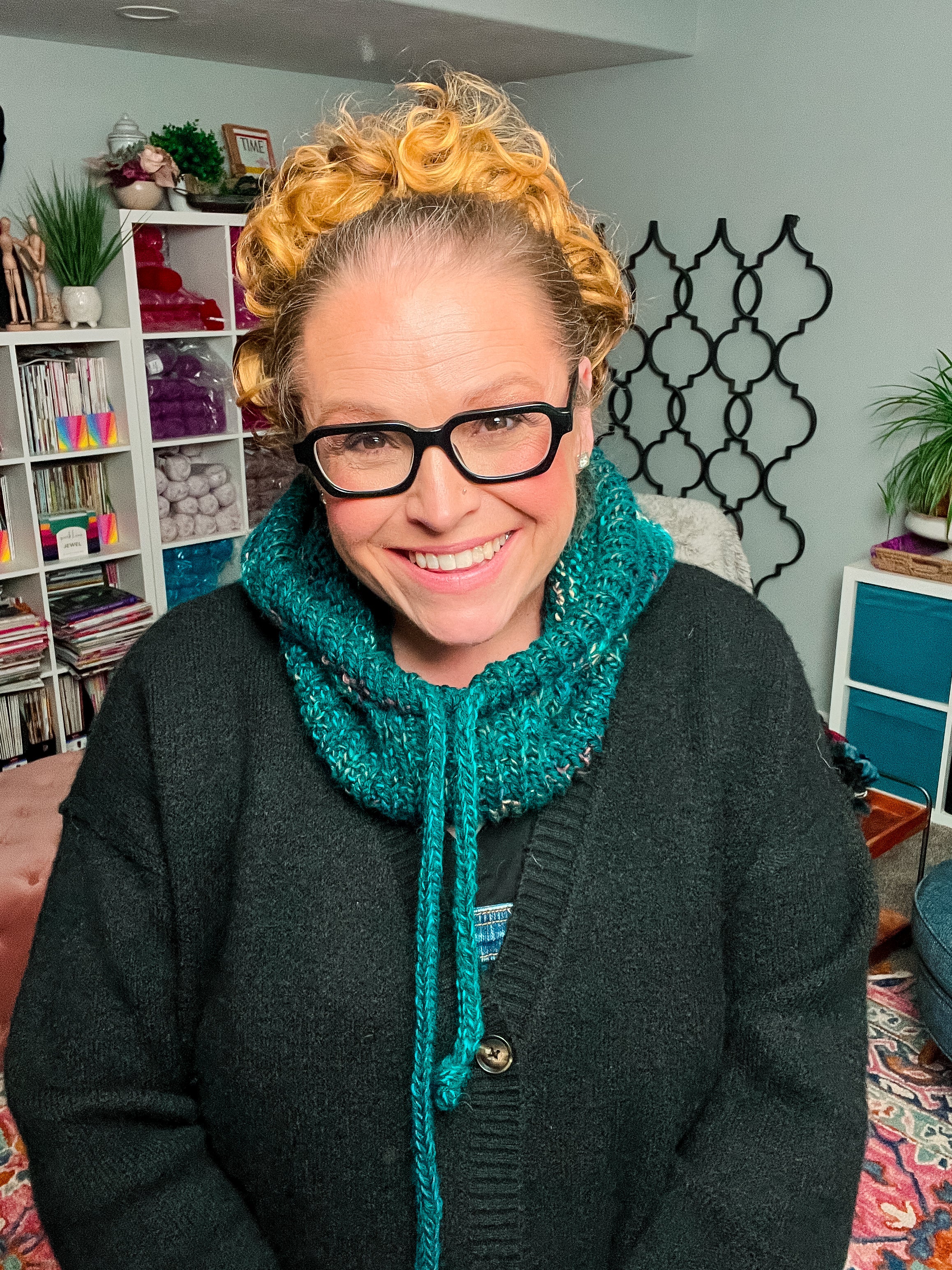 Sure! Here is the revised sentence using your product data: A smiling woman with curly hair and glasses wears a teal Cozy Sideline Knit Balaclava, made from worsted weight yarn and styled with a black sweater, standing indoors amid colorful shelves stocked with yarn and surrounded by plants in the background.