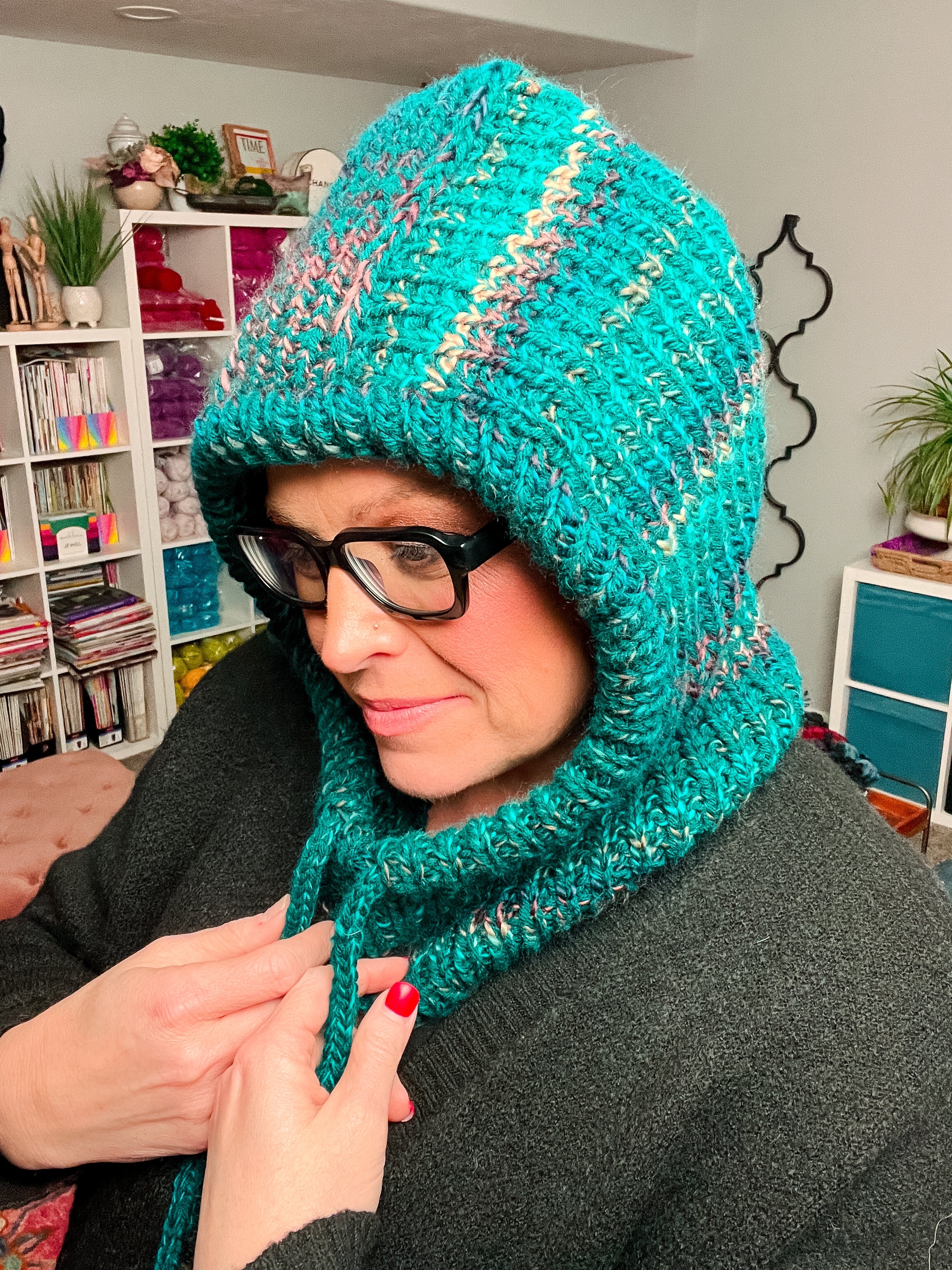A person wearing glasses and a dark sweater is smiling while showcasing the Cozy Sideline Knit Balaclava – PDF, a cozy hooded cowl pattern made with worsted weight yarn. The teal balaclava features ties that the person is holding, and shelves lined with yarn and books are visible in the background.