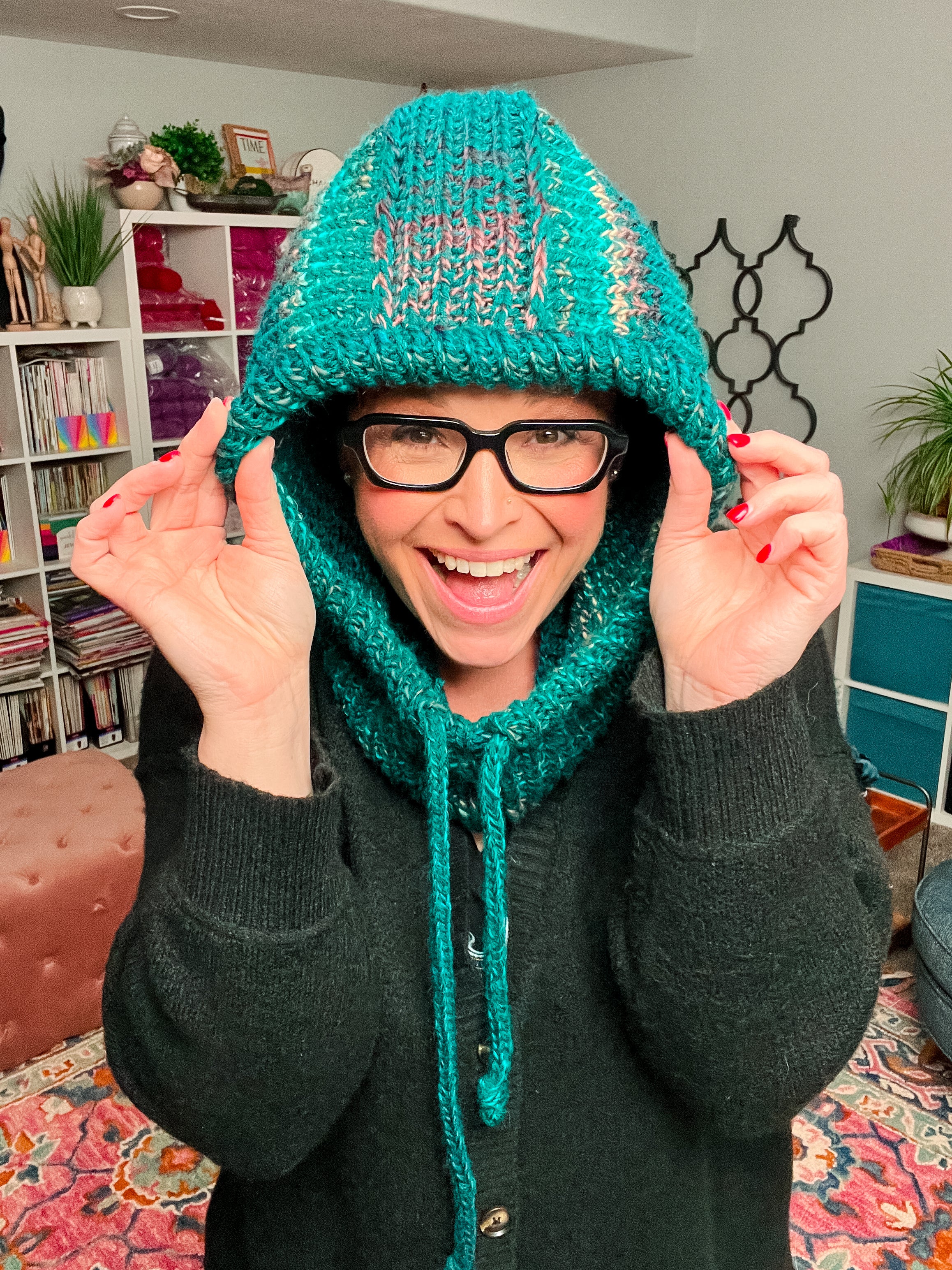 A smiling person with glasses wears the Cozy Sideline Knit Balaclava—crafted from worsted weight yarn in a teal shade—holding the sides with both hands. Shelves, plants, and colorful decor appear in the background.