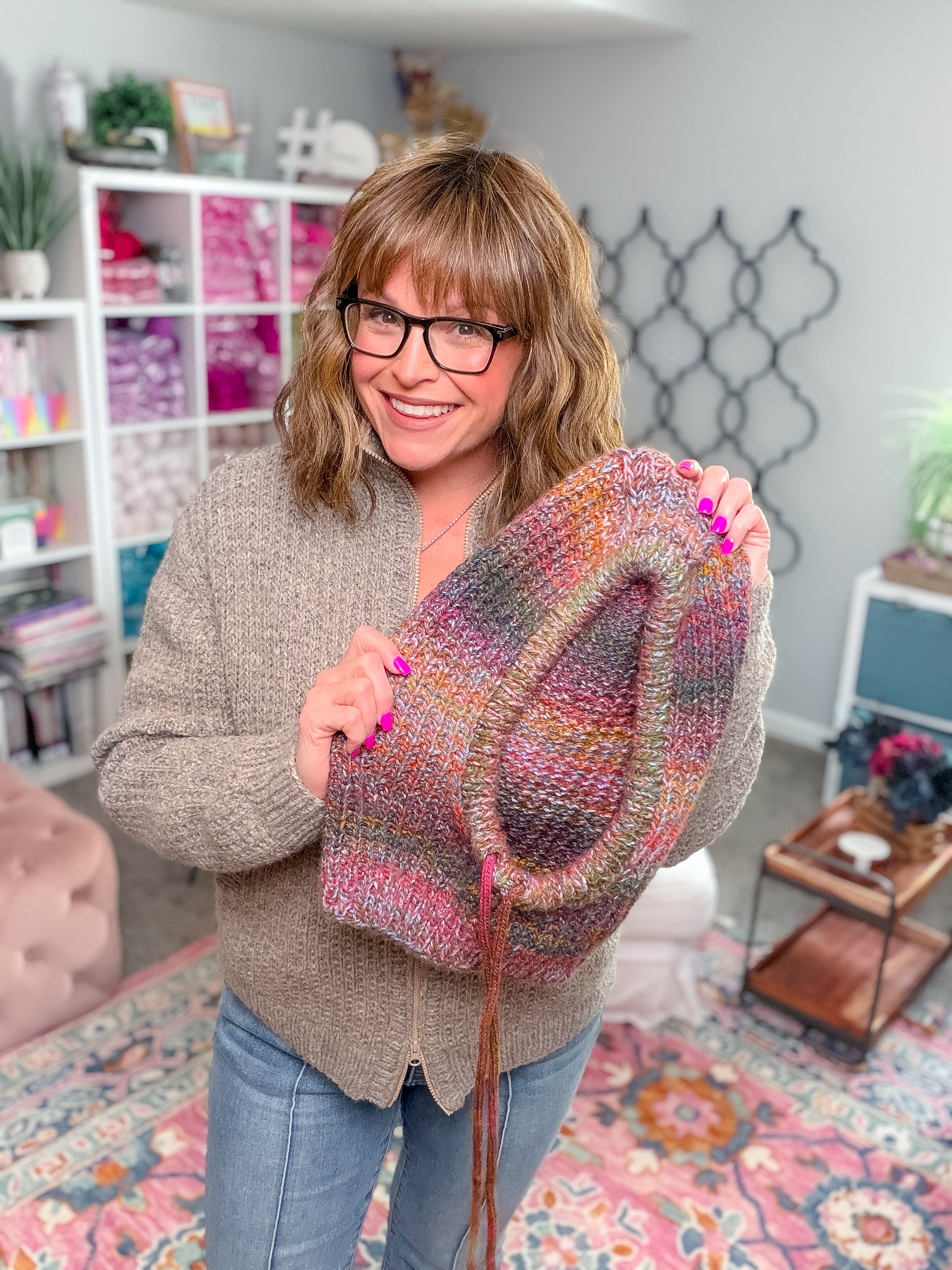 A smiling woman holds up a multicolored Cozy Sideline Knit Balaclava - PDF made with worsted weight yarn.