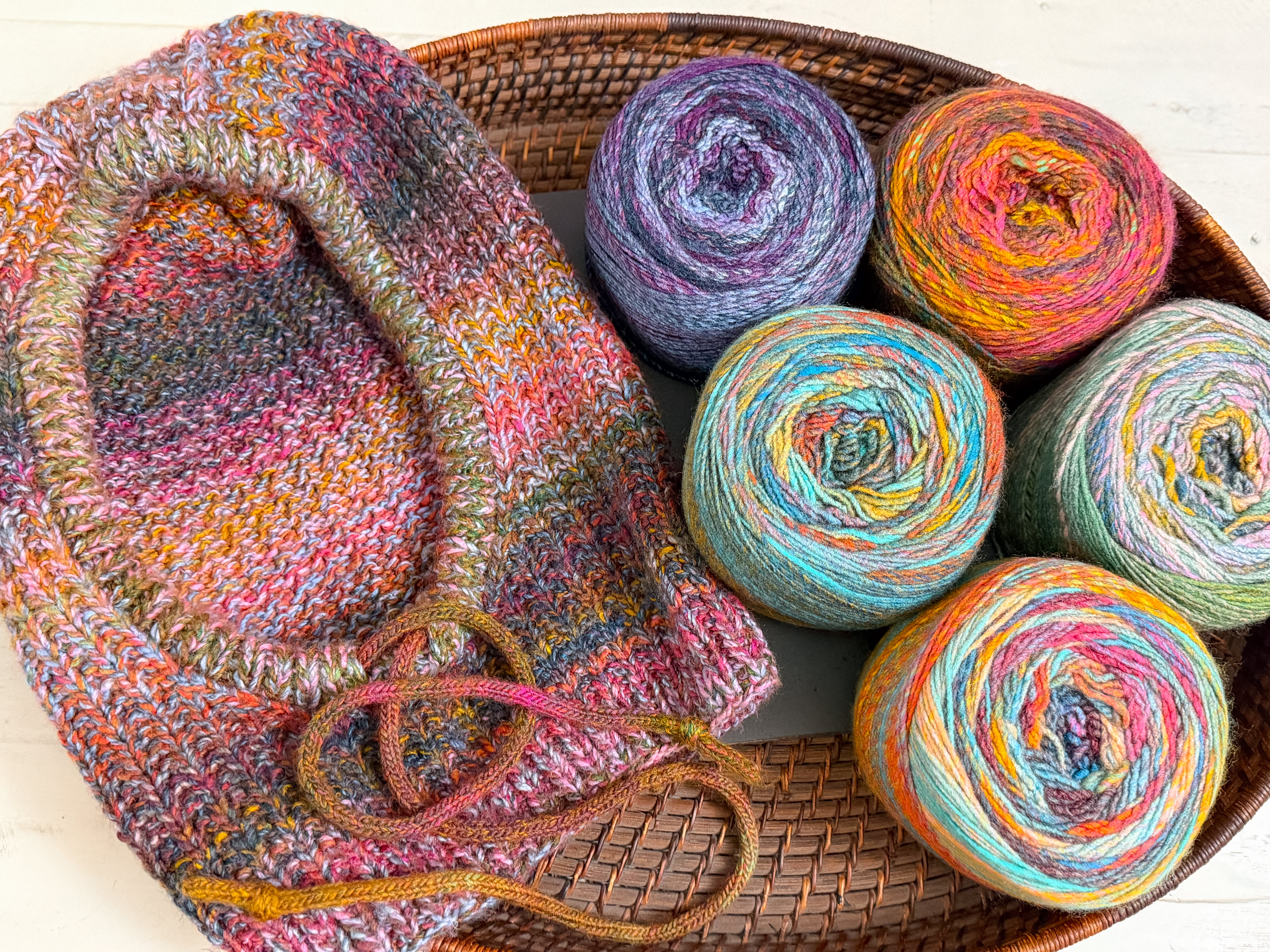A Cozy Sideline Knit Balaclava - PDF sits in a round wicker basket next to five colorful cakes of worsted weight yarn.
