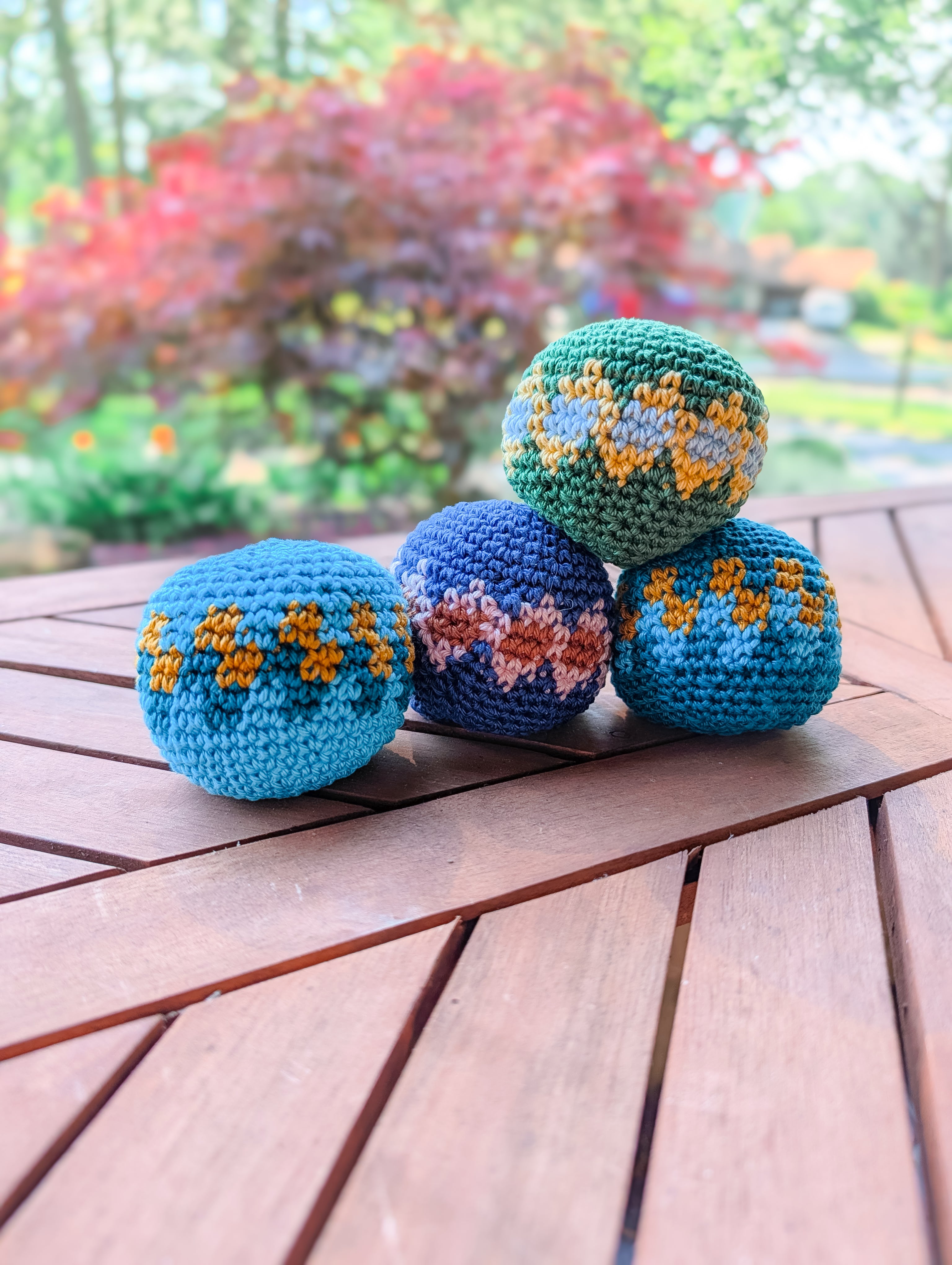 Four vibrant crochet balls made with the Crochet Hacky Sack Pattern – Beginner Friendly Crochet Ball are stacked in a pyramid on a wooden table outdoors, perfect for anyone seeking an easy, quick stash busting project. Lush greenery blurs in the background.
