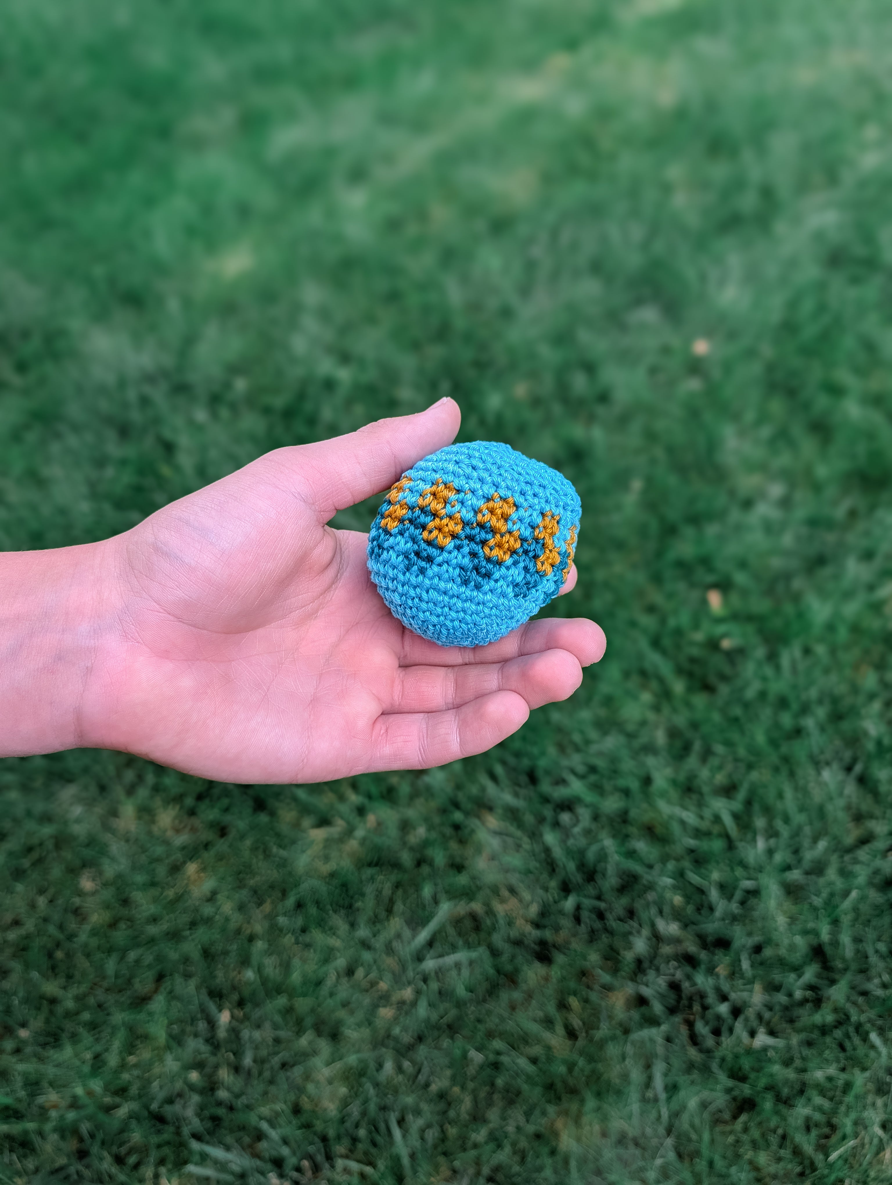 A hand holds a small, blue ball made with the “Crochet Hacky Sack Pattern – Beginner Friendly Crochet Ball (Quick Stash Busting Project)” featuring orange and yellow details. The background is blurred green grass.