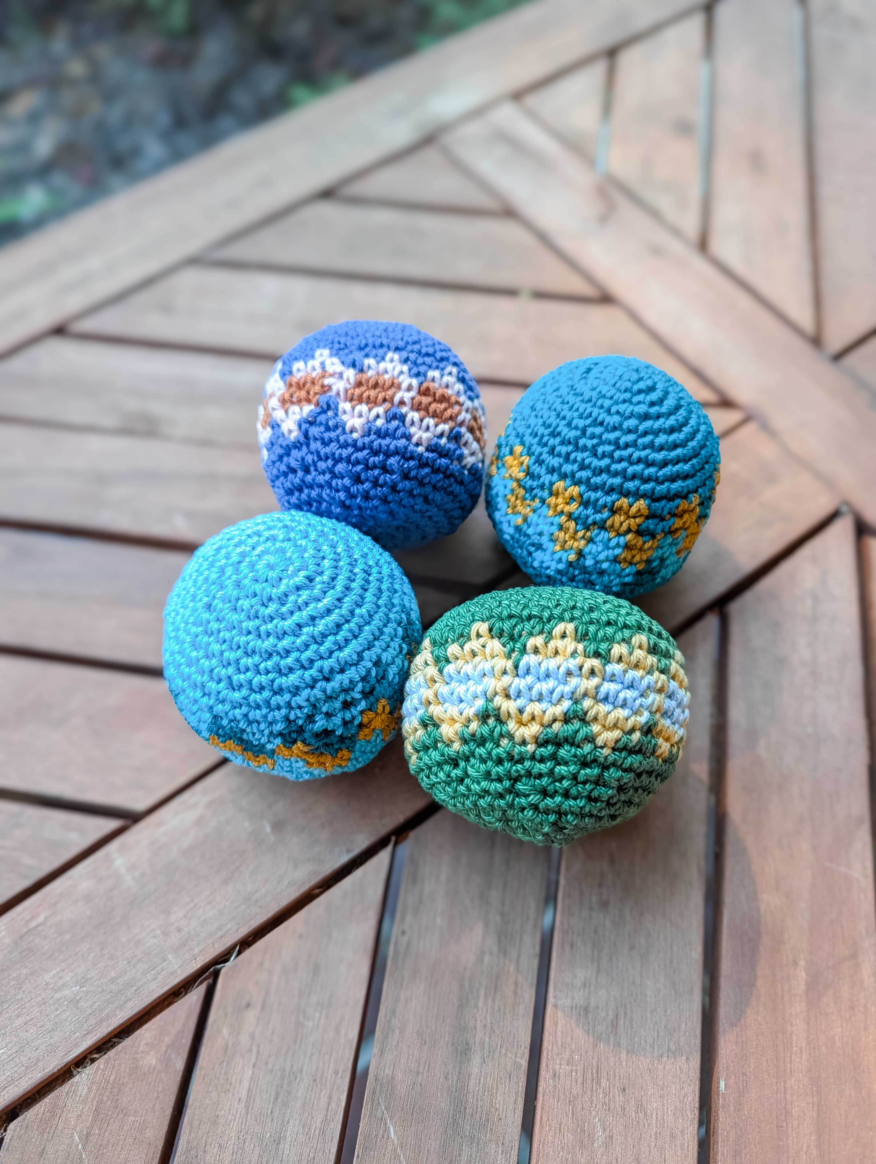 Four crochet balls with blue, green, yellow, brown, and white tapestry patterns rest on a wooden table—made from the Crochet Hacky Sack Pattern – Beginner Friendly Crochet Ball (Quick Stash Busting Project). Perfect for beginners or as playful hacky sacks.