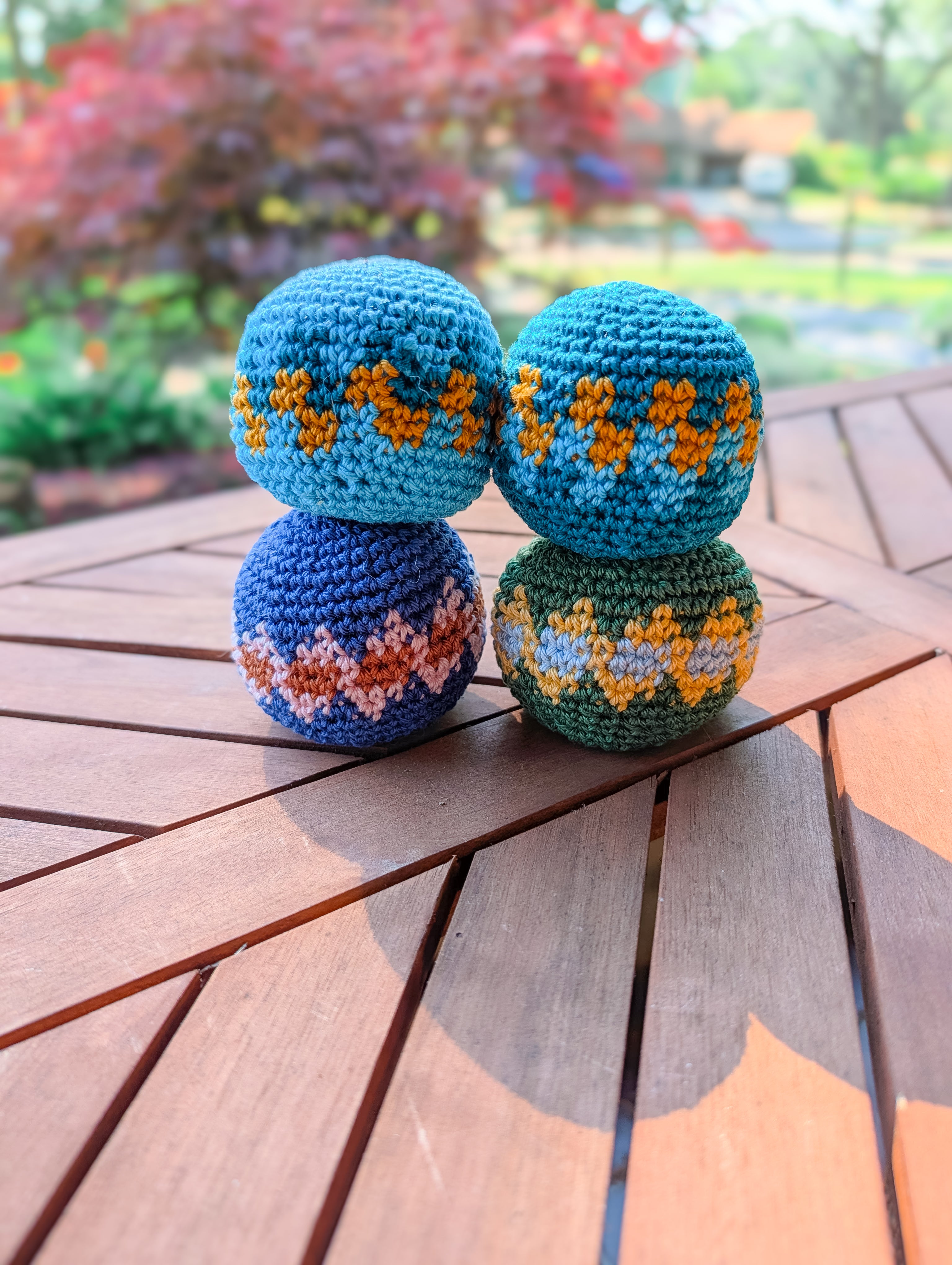 Four colorful crochet hacky sacks are stacked on a wooden table outdoors with greenery and a red-leaved tree in the background—made using the Crochet Hacky Sack Pattern – Beginner Friendly Crochet Ball (Quick Stash Busting Project).