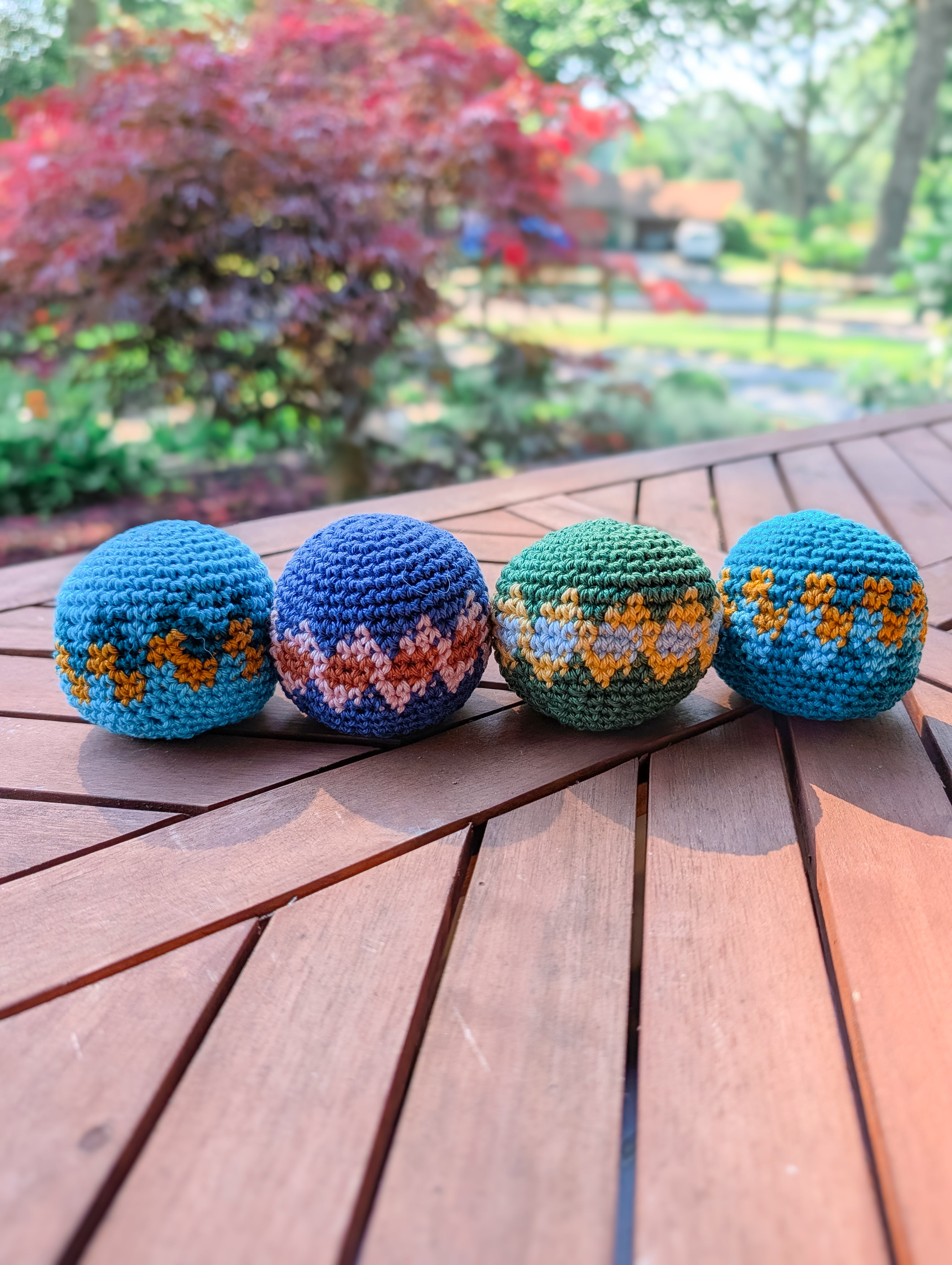 Four beginner-friendly Crochet Hacky Sack Balls made with the Crochet Hacky Sack Pattern – Beginner Friendly Crochet Ball rest in blue, green, and multicolored designs on a wooden outdoor table, garden softly blurred in the background.