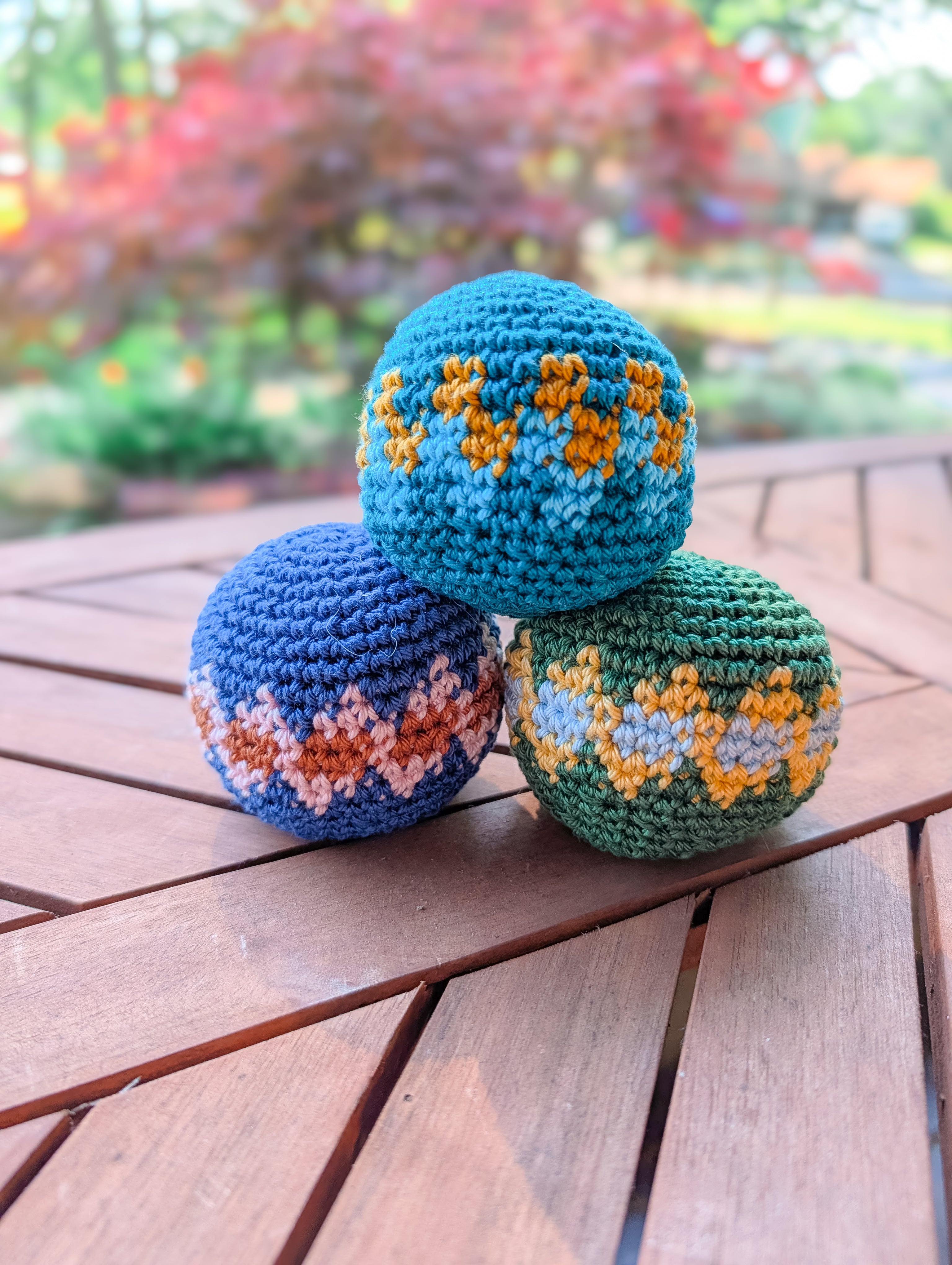 Three geometric crocheted balls in blue, green, and teal yarn are stacked on a wooden table outdoors. Made with the Crochet Hacky Sack Pattern – Beginner Friendly Crochet Ball (Quick Stash Busting Project). Perfect for beginners!.