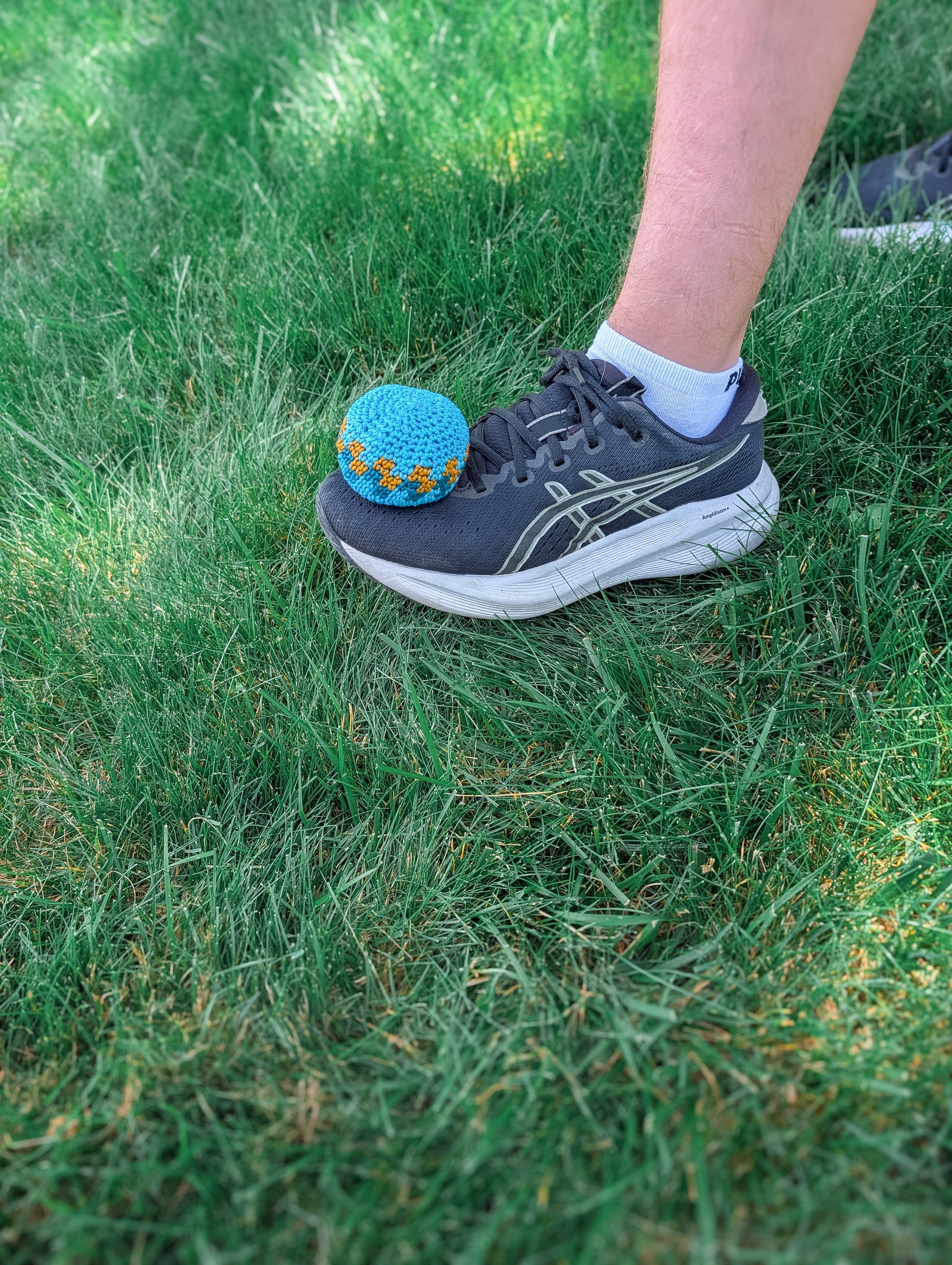 A person wearing a gray sneaker steps on a handmade ball created from the Crochet Hacky Sack Pattern – Beginner Friendly Crochet Ball, while standing on green grass. Only the lower leg and foot are visible.