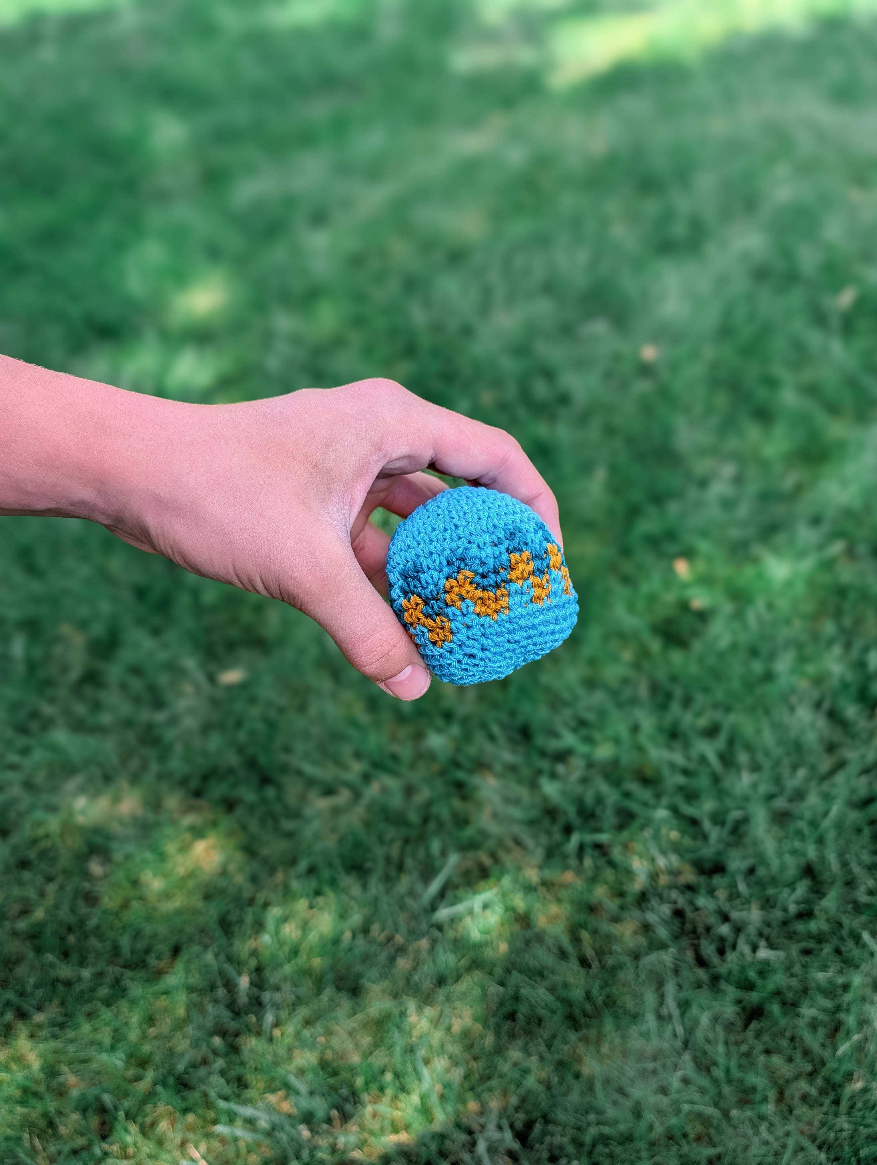A hand holds a small, blue crochet ball featuring an orange zigzag, made using the Beginner-Friendly Crochet Hacky Sack Pattern—a quick stash-busting project—against a blurred green grass background.