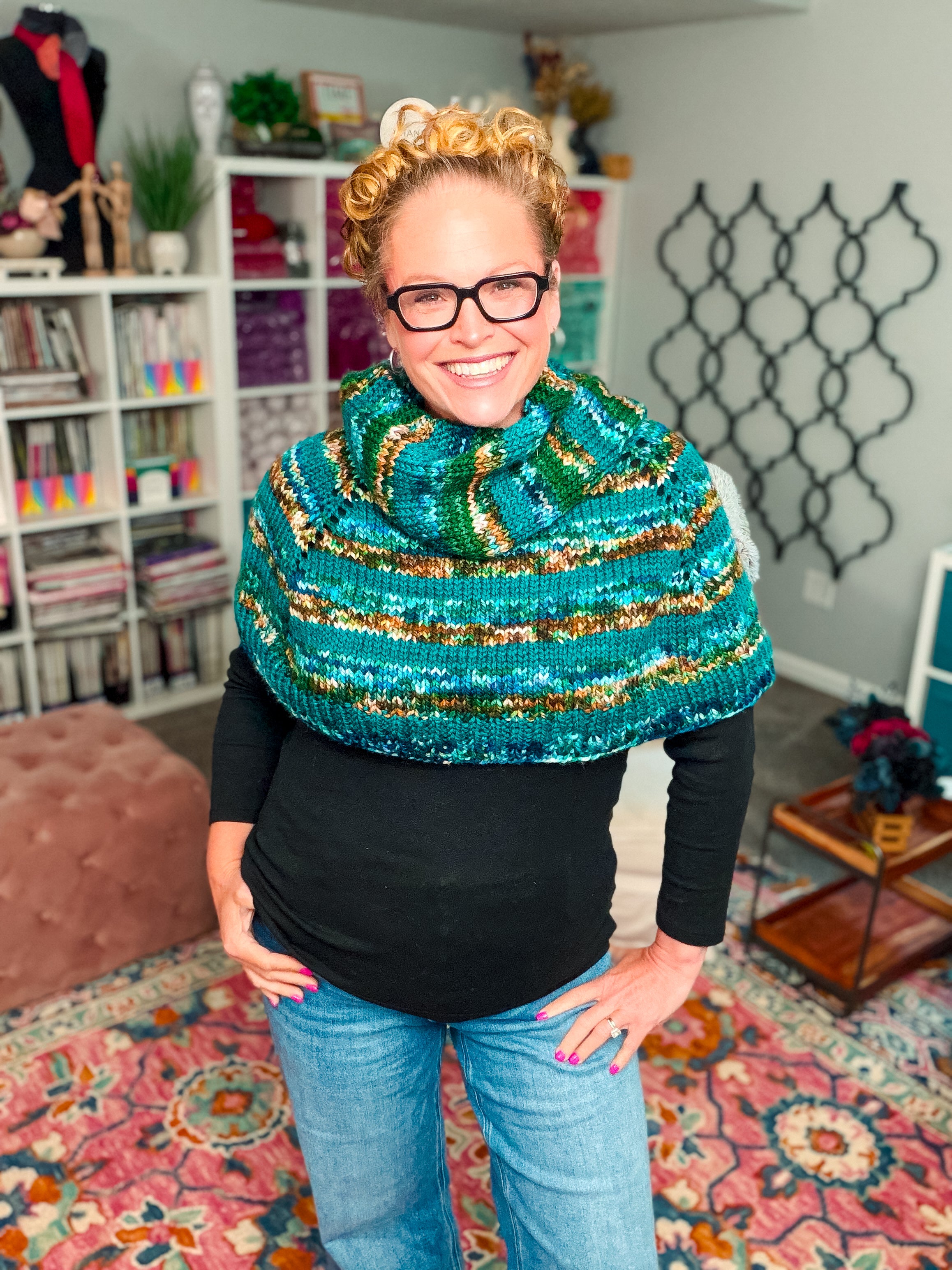 A smiling person with glasses and curly hair wears the Deschutes Knit Capelet, a chunky, seamless shoulder wrap featuring jogless stripes in green and blue, layered over a black top in a cozy, colorful room lined with books and yarn.