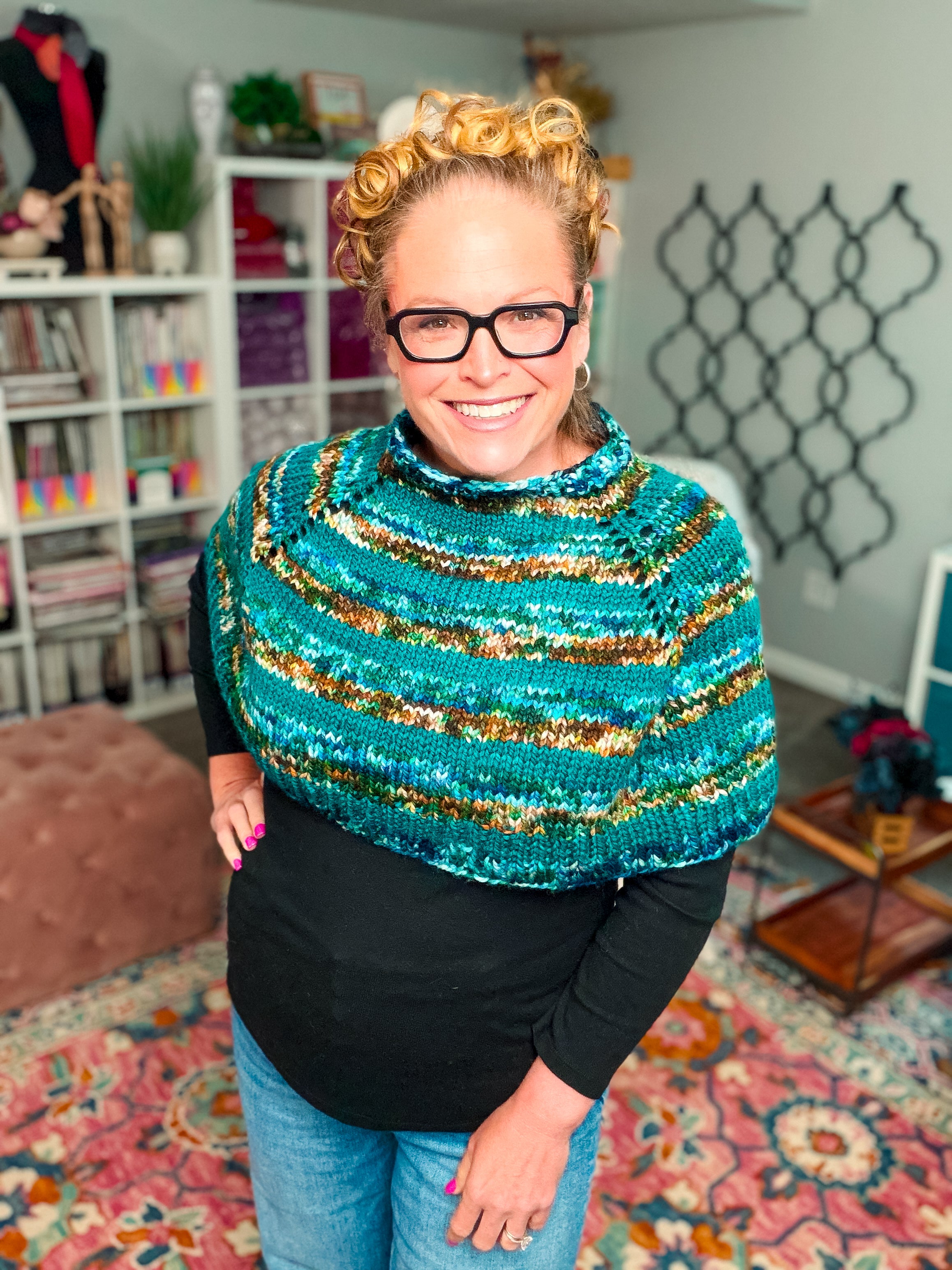A smiling person with curly hair and glasses wears the Deschutes Knit Capelet—a chunky, blue, green, and brown shoulder wrap crafted using the jogless stripe technique—over a black top, standing in a cozy, colorful room filled with bookshelves, yarn, and a patterned rug.
