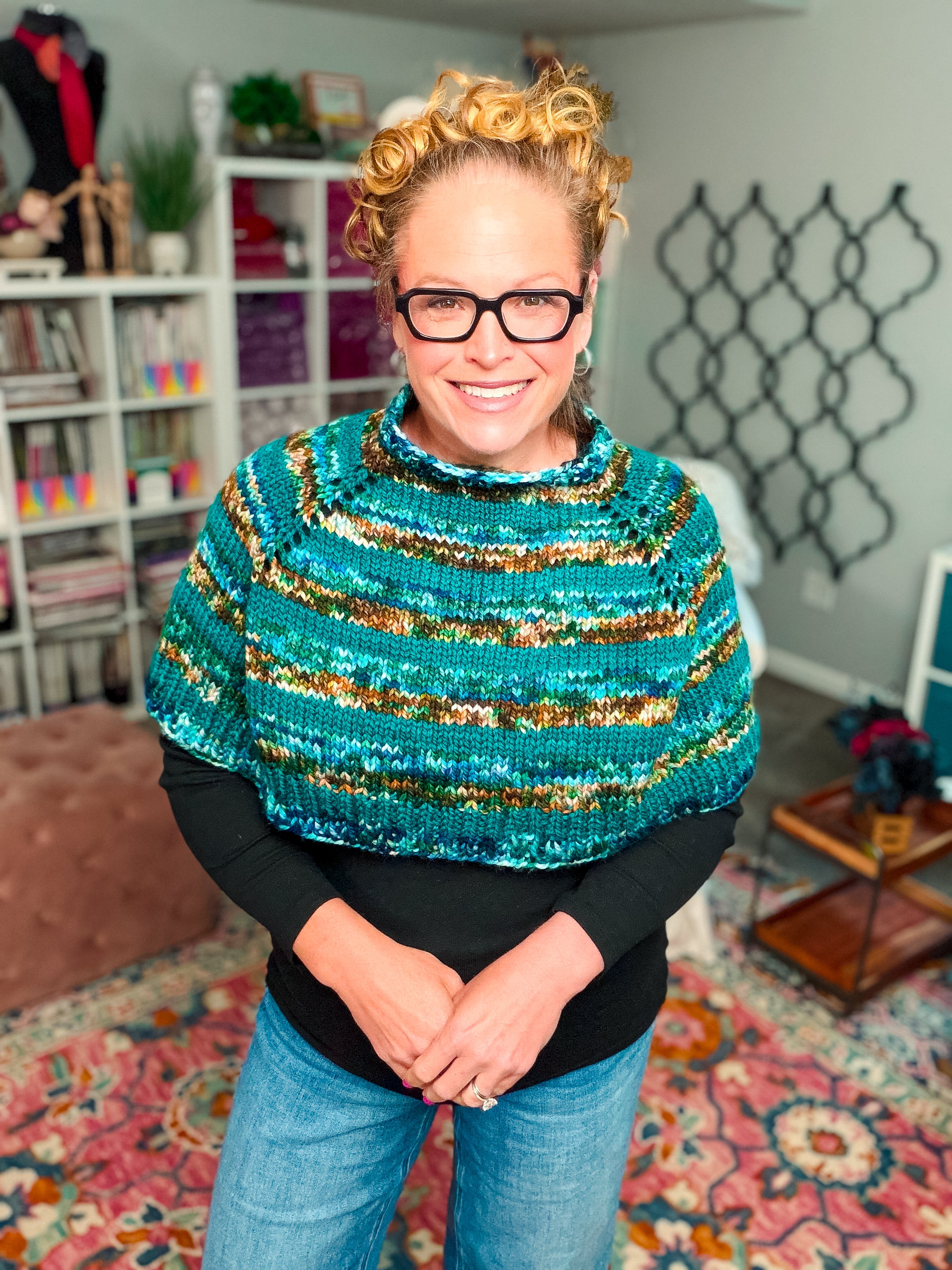 A smiling woman with curly hair and glasses wears the Deschutes Knit Capelet, a chunky knit shoulder wrap featuring expert jogless stripes in blue and green, standing in a colorful, cozy room with bookshelves and a patterned rug.